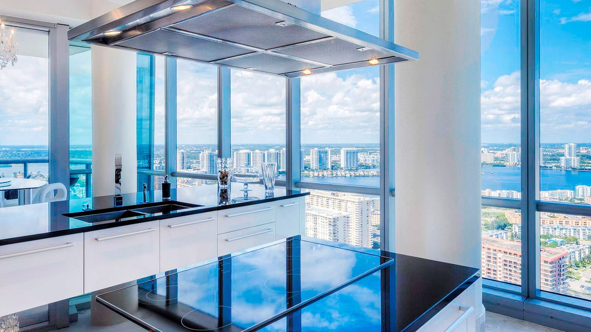 Corner chef kitchen inside one of the luxury and ultra luxury condos at Jade Ocean in Sunny Isles Beach, with glossy black counters, floor-to-ceiling glass, and expansive skyline and bay views.