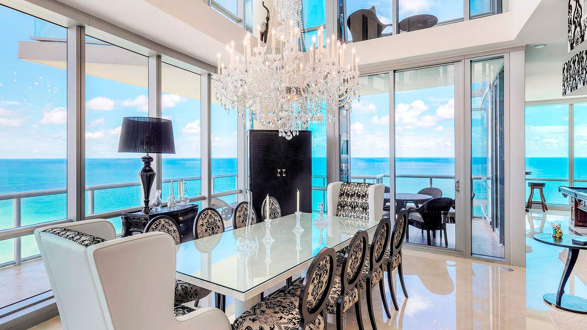 Double-height dining room inside one of the luxury and ultra luxury condos at Jade Ocean in Sunny Isles Beach, with a crystal chandelier, patterned chairs, walls of glass, and an oceanfront balcony.