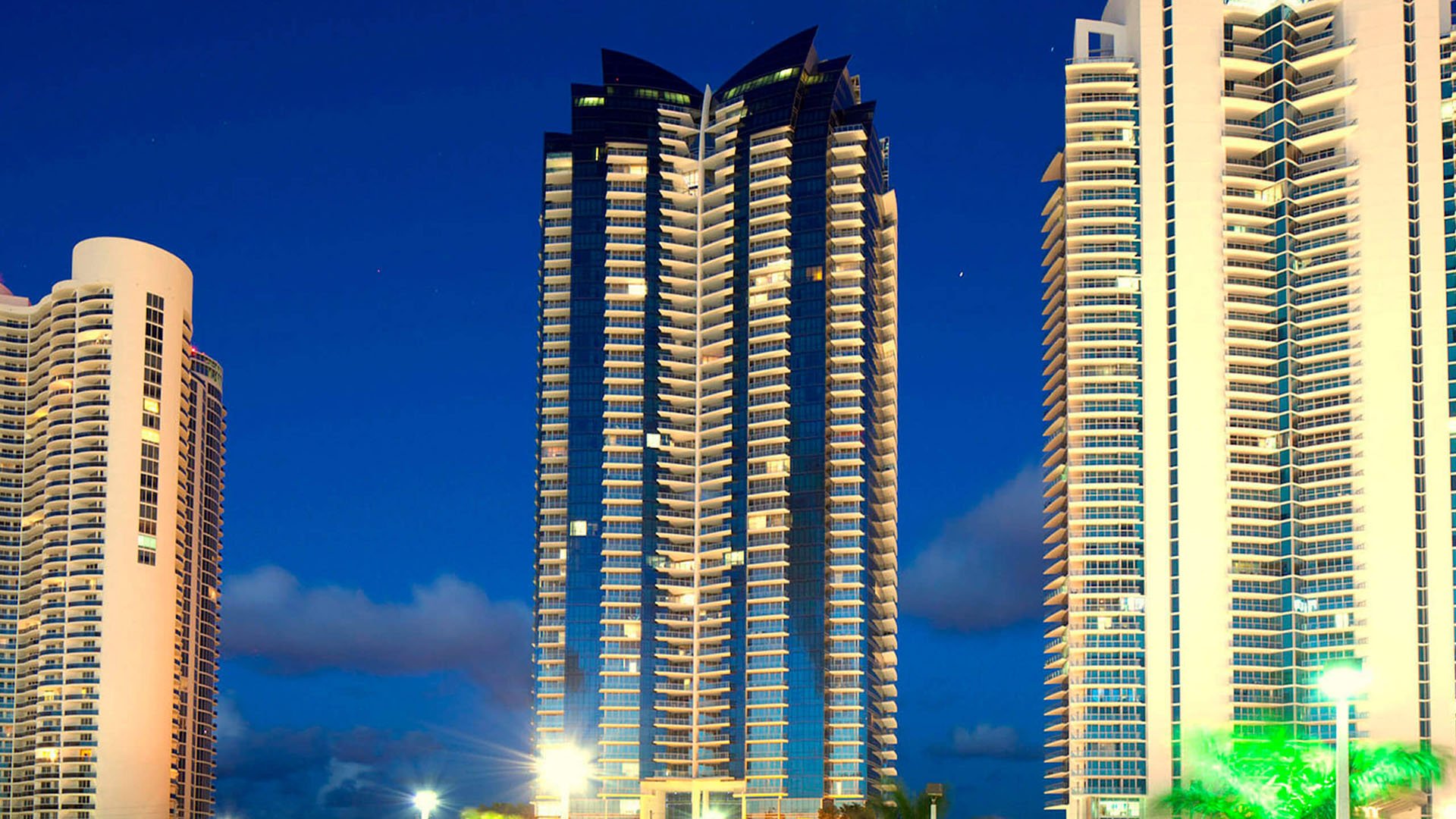 Illuminated night exterior of Jade Ocean in Sunny Isles Beach, showcasing luxury and ultra luxury condos with the signature glass tower glowing between neighboring beachfront high-rises.