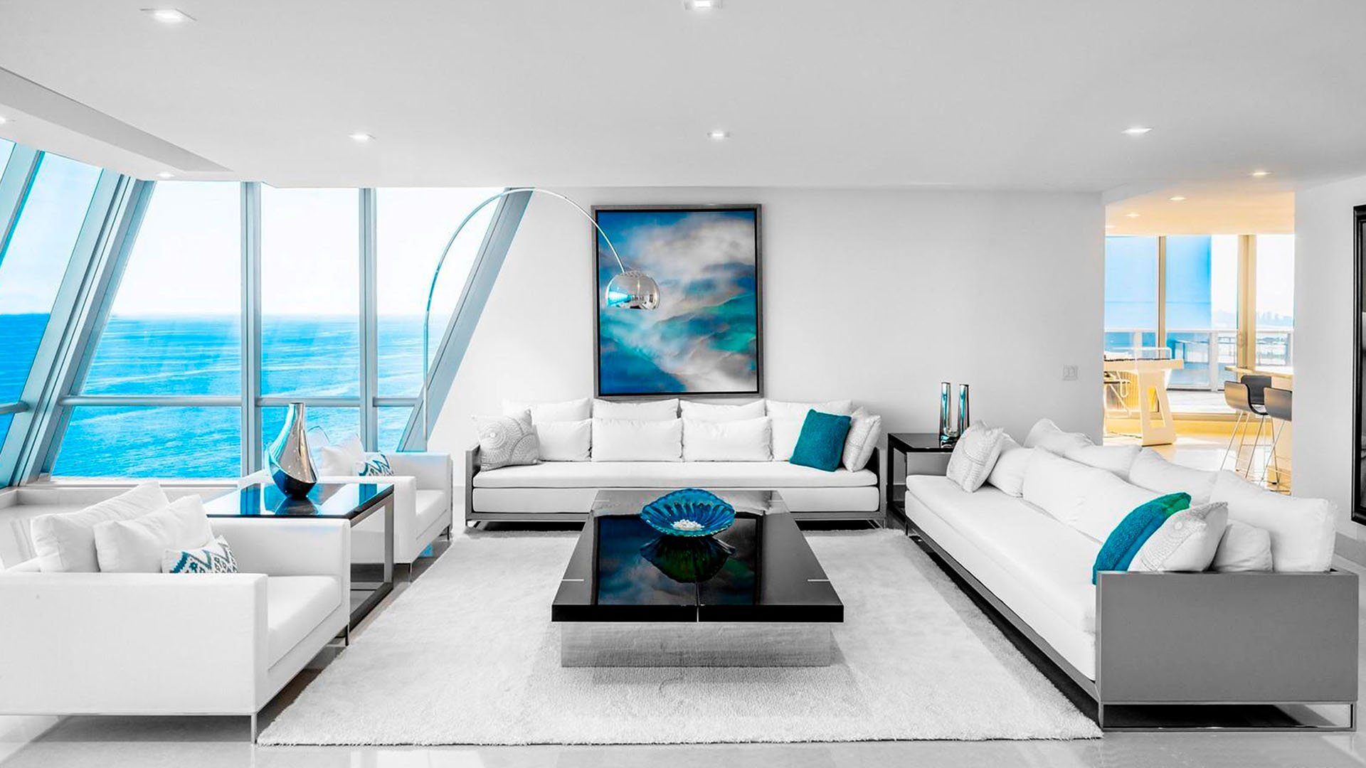 Minimalist oceanfront living room inside one of the luxury and ultra luxury condos at Jade Ocean in Sunny Isles Beach, featuring white seating, a dramatic angled glass wall, blue accents, and open water views.