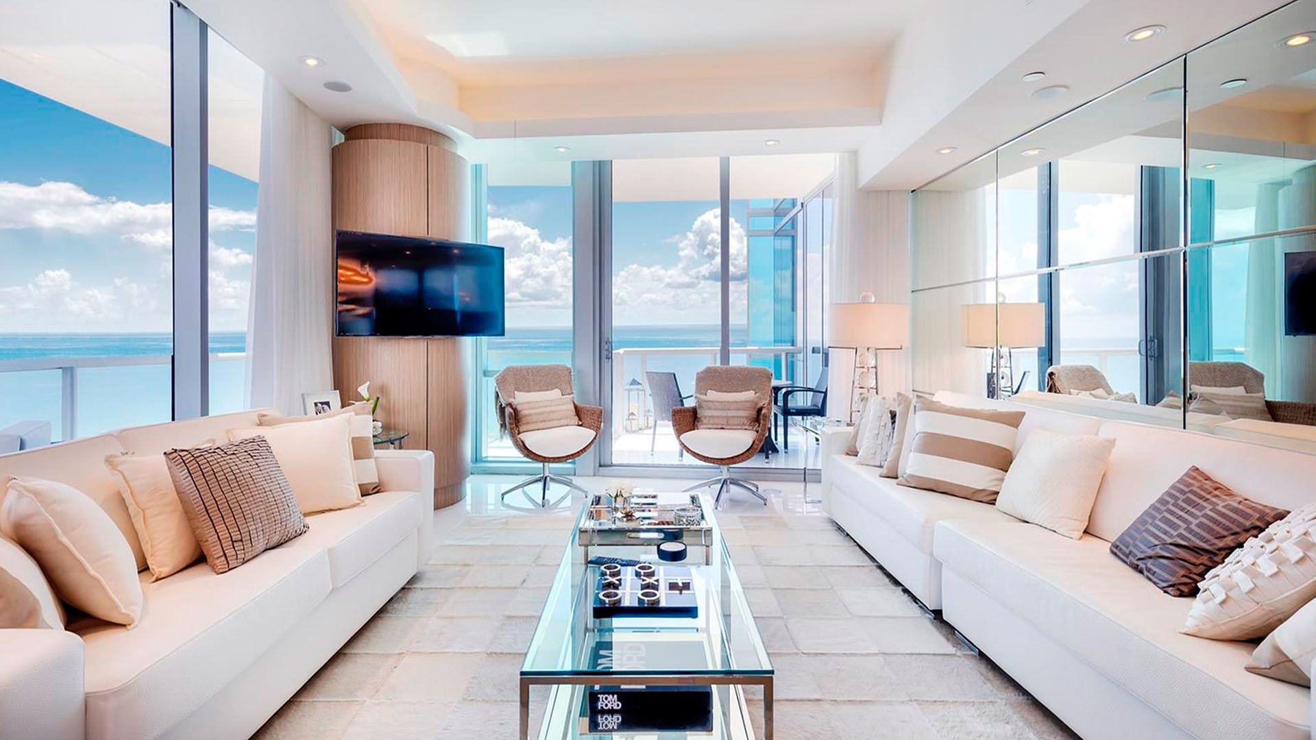 Oceanfront living room inside one of the luxury and ultra luxury condos at Jade Ocean in Sunny Isles Beach, with mirrored walls, white sofas, a glass coffee table, and a balcony facing the water.