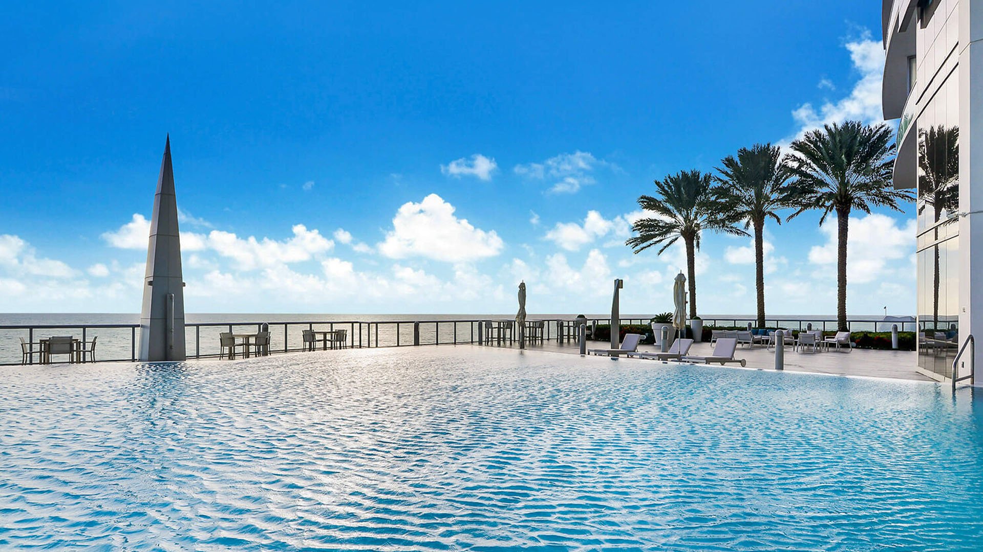 Oceanfront pool terrace at Jade Ocean in Sunny Isles Beach, highlighting luxury and ultra luxury condos with a rippling resort pool, obelisk sculpture, palm trees, and unobstructed Atlantic views.