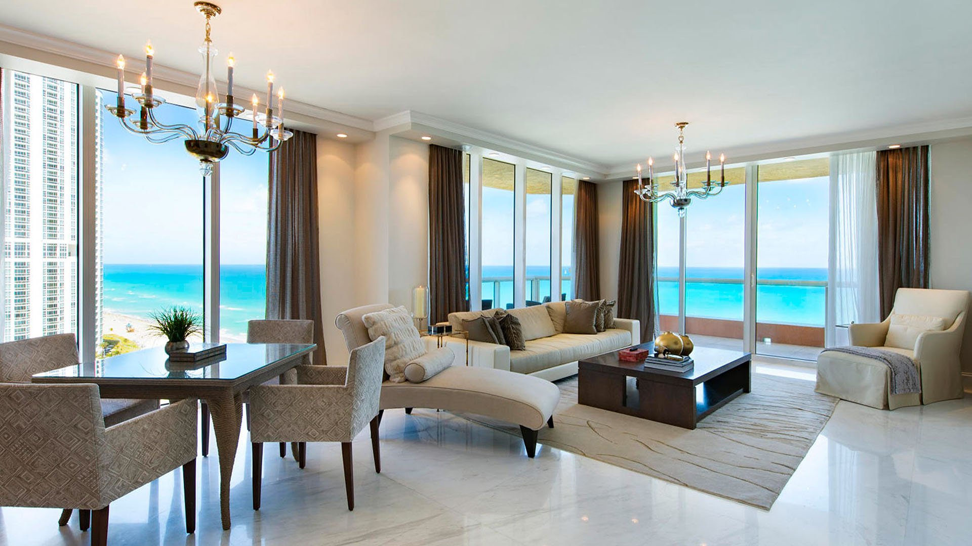 Open-concept living and dining room inside one of the luxury and ultra luxury condos at Jade Ocean in Sunny Isles Beach, with wraparound glass, a corner balcony, chandeliers, and bright ocean views.