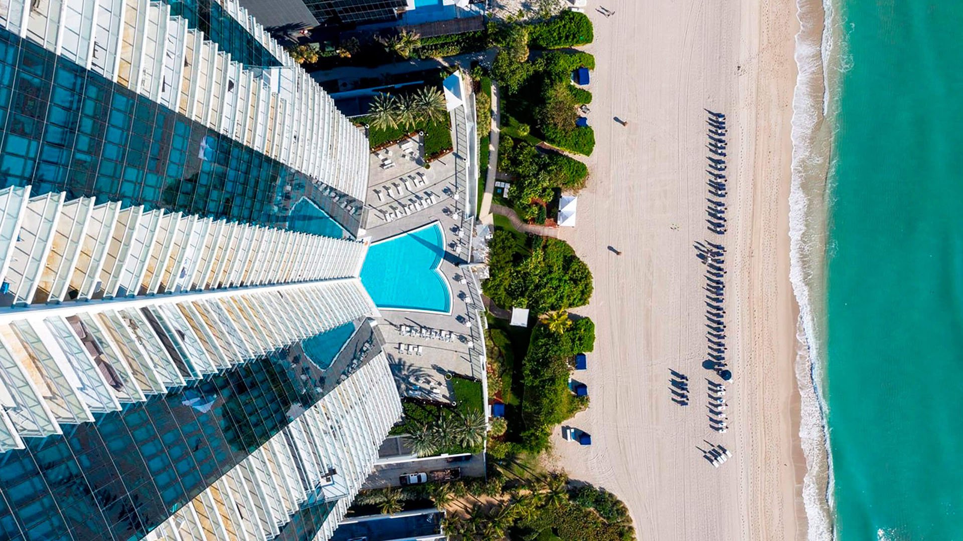 Top-down aerial of Jade Ocean in Sunny Isles Beach, revealing luxury and ultra luxury condos with the beachfront pool deck, landscaped grounds, and sandy shoreline meeting turquoise water.
