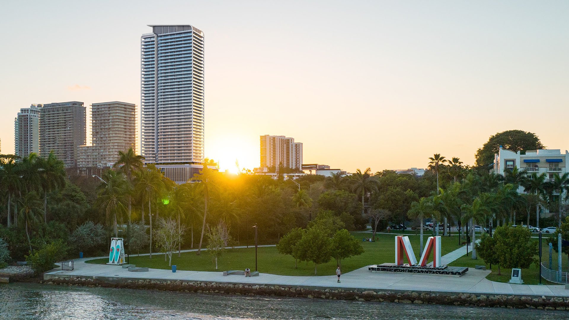 Sunset view of Jean-Georges Miami Tropic Residences near a waterfront park on Biscayne Bay, Miami, Florida, with skyline backdrop and bayfront greenery, positioning luxury and ultra luxury preconstruction condos in a prime location.