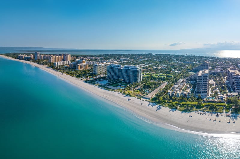Key Biscayne luxury and ultra luxury condos in an aerial beachfront view with a curved sandy shoreline, turquoise water, oceanfront residences, and a lush island neighborhood stretching to the horizon.