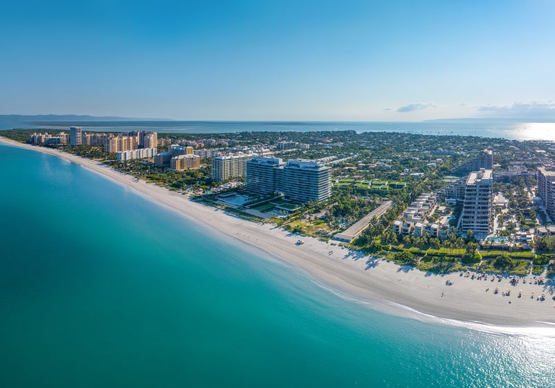 Key Biscayne luxury and ultra luxury condos in an aerial beachfront view with a curved sandy shoreline, turquoise water, oceanfront residences, and a lush island neighborhood stretching to the horizon.