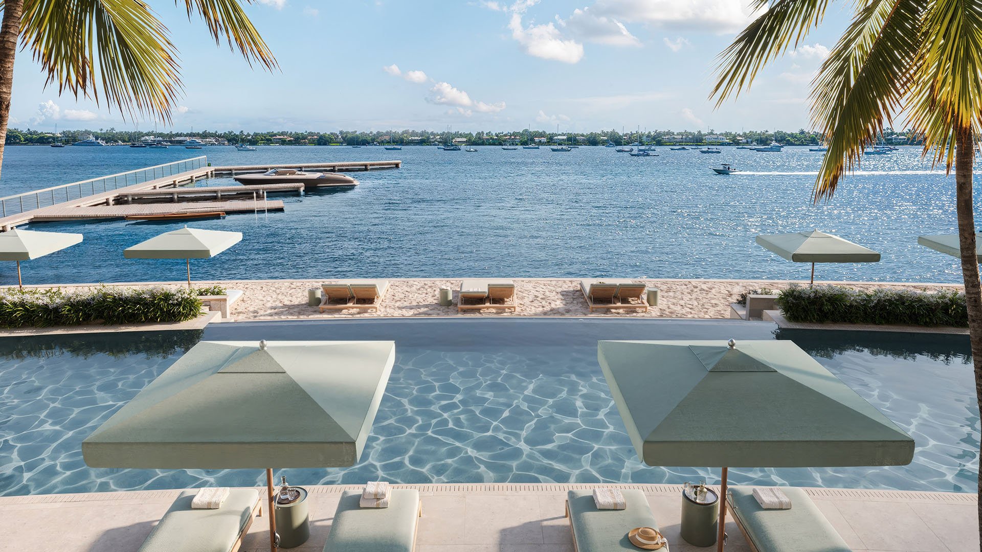 Poolside umbrellas and sandy loungers overlook a dock and open waterfront at Mandarin Oriental Residences, West Palm Beach, ultra luxury condos in West Palm Beach with a luxury resort-style water view.