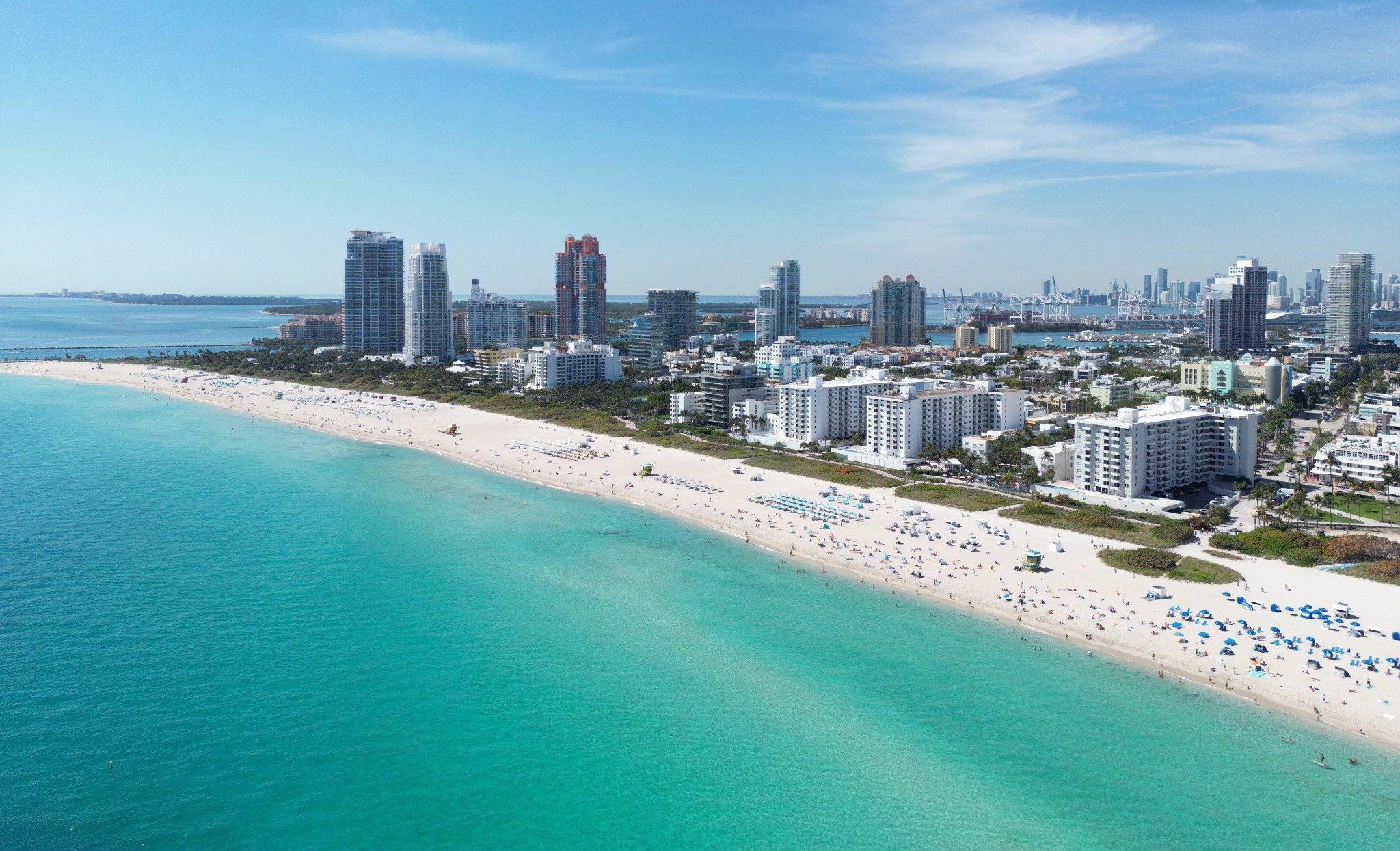 Miami Beach luxury and ultra luxury condos in an aerial beachfront skyline view with turquoise water, wide white sand, oceanfront towers, and the city skyline with port cranes in the distance.