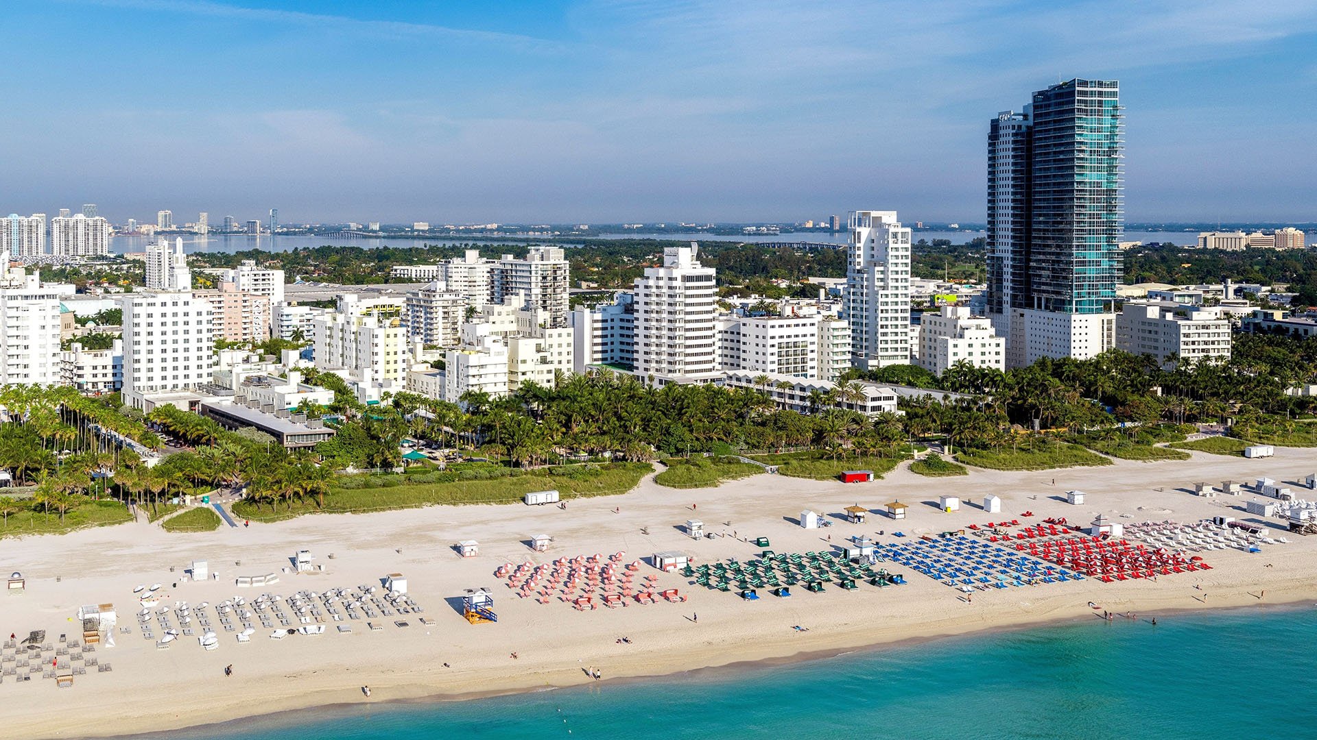 Mila Bay Harbor Islands preconstruction luxury and ultra luxury condos in Bay Harbor Islands near a beachfront skyline view with turquoise water, sandy shoreline, resort towers, and rows of colorful umbrellas.