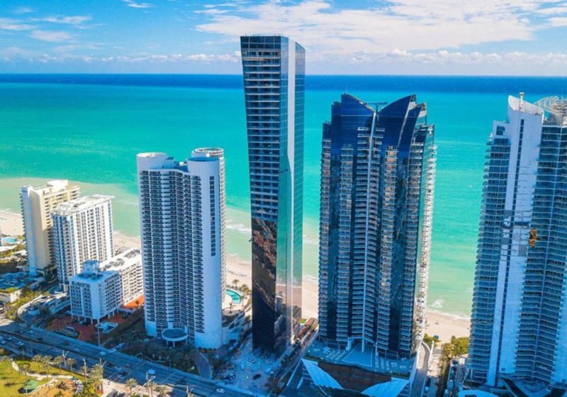 Aerial beachfront view of Muse Residences in Sunny Isles Beach beside turquoise water and neighboring towers, highlighting luxury and ultra luxury condos along the oceanfront.