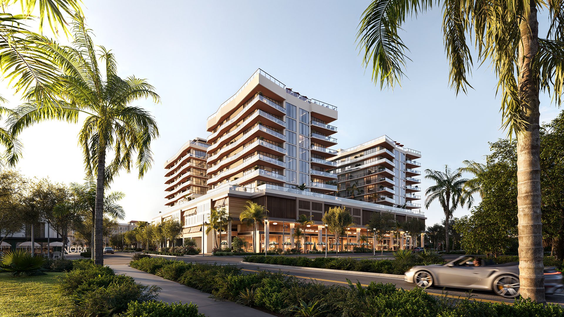 Angled streetfront view of Nora House in West Palm Beach, luxury and ultra luxury condos with stepped glass balconies, palm-lined landscaping, glowing retail frontage, and a tropical boulevard setting.