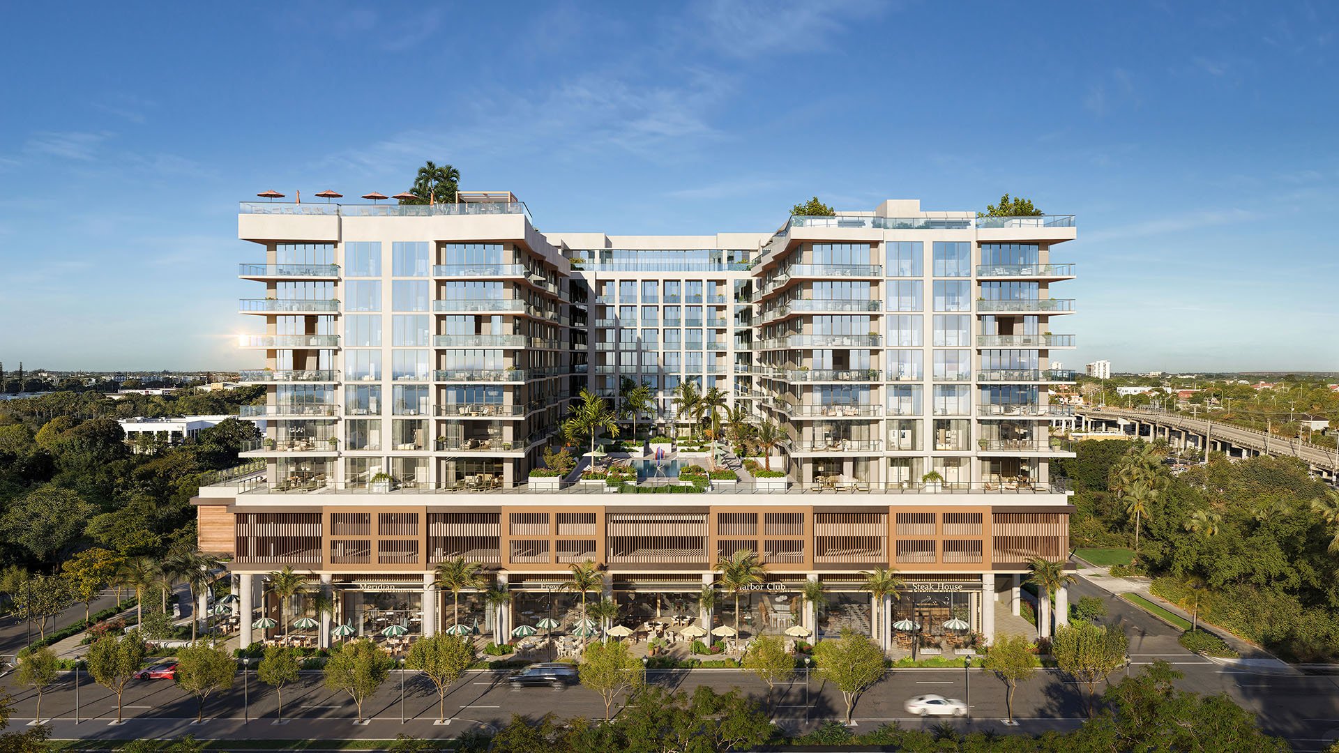 Daylight front elevation of Nora House in West Palm Beach, luxury and ultra luxury condos showing the full glass facade, elevated courtyard pool, rooftop terraces, street trees, and ground-floor retail along the avenue.