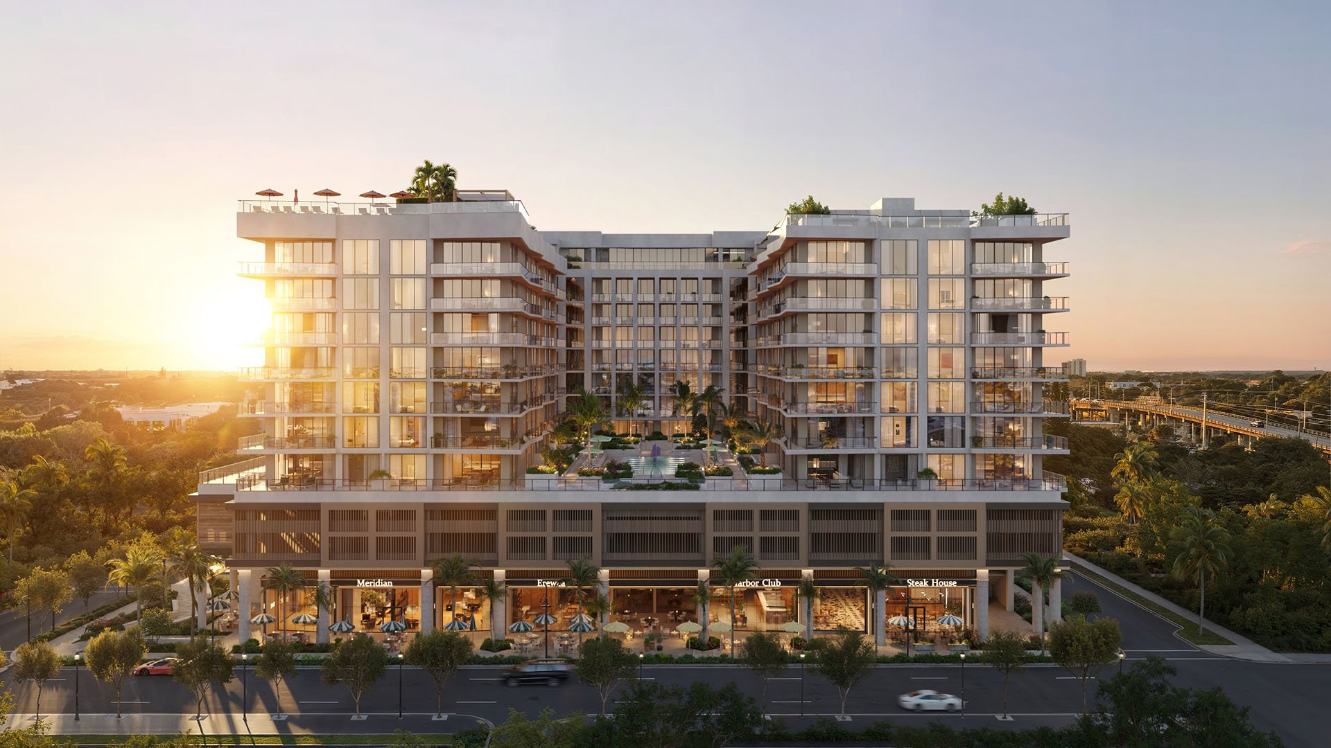 Sunset front elevation of Nora House in West Palm Beach, luxury and ultra luxury condos highlighting the illuminated glass facade, elevated courtyard pool, rooftop terraces, tree-lined street, and the ground-floor retail podium.