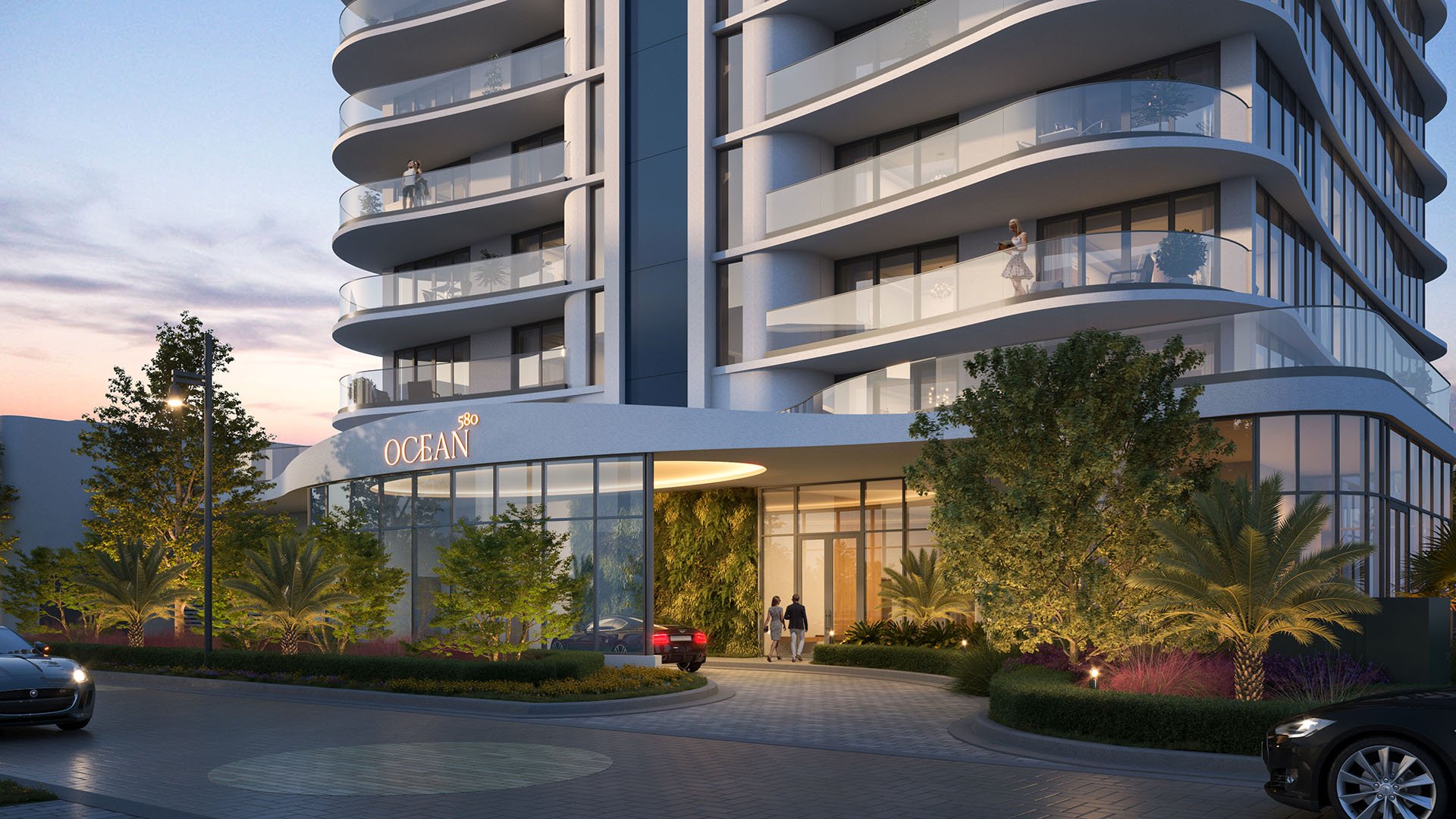 Dusk entrance at Ocean 580 in Pompano Beach, preconstruction luxury and ultra luxury condos with illuminated glazing, curved balconies, a porte cochere arrival court and tropical landscaping.