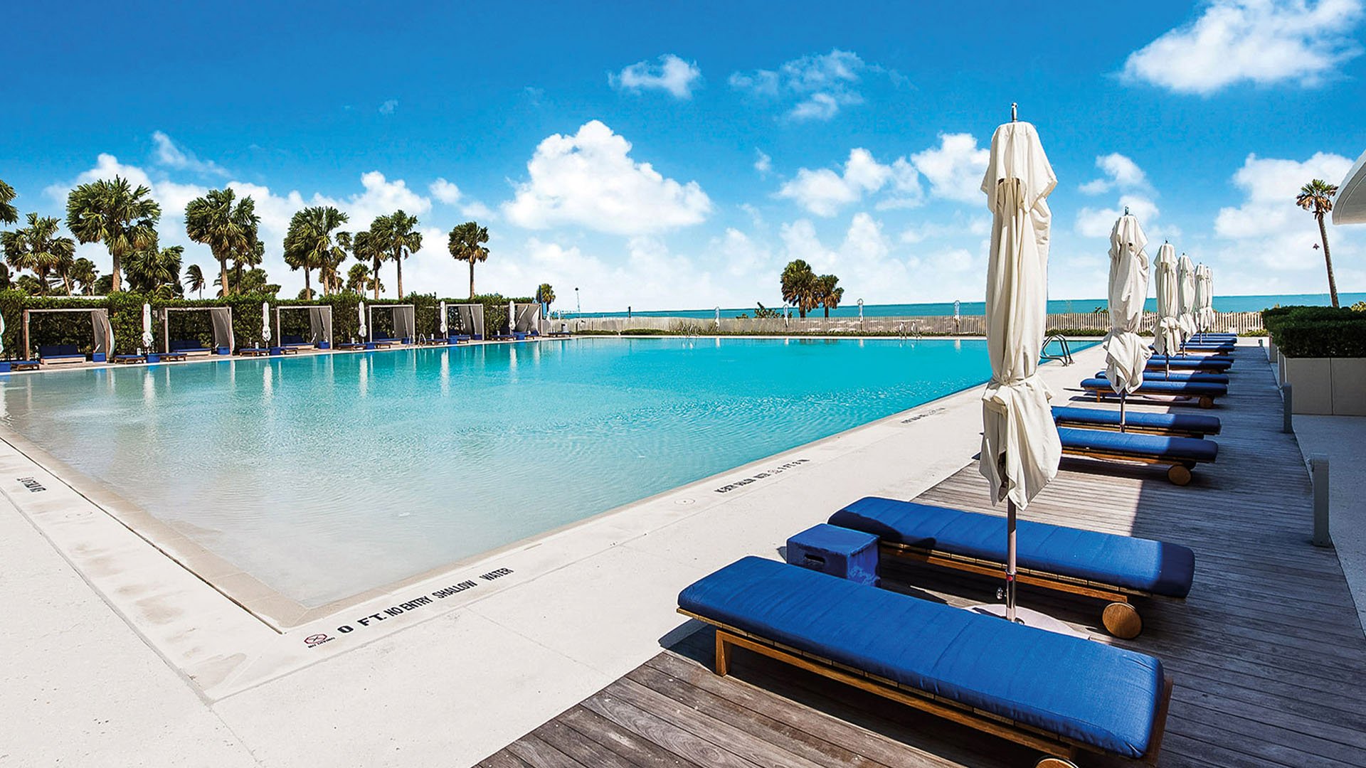 Oceanfront pool deck at Oceana Key Biscayne in Key Biscayne, where luxury and ultra luxury condos overlook a large resort pool, blue loungers, shaded cabanas, palms, and the beach.