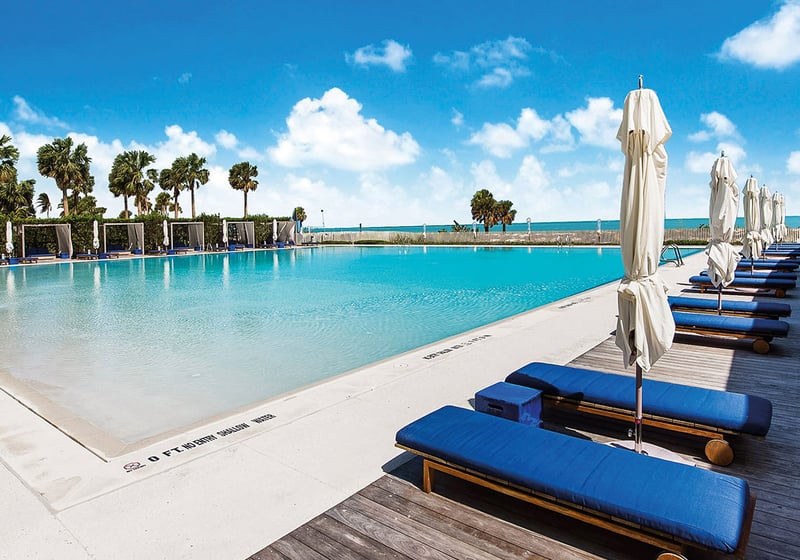 Oceanfront pool deck at Oceana Key Biscayne in Key Biscayne, where luxury and ultra luxury condos overlook a large resort pool, blue loungers, shaded cabanas, palms, and the beach.
