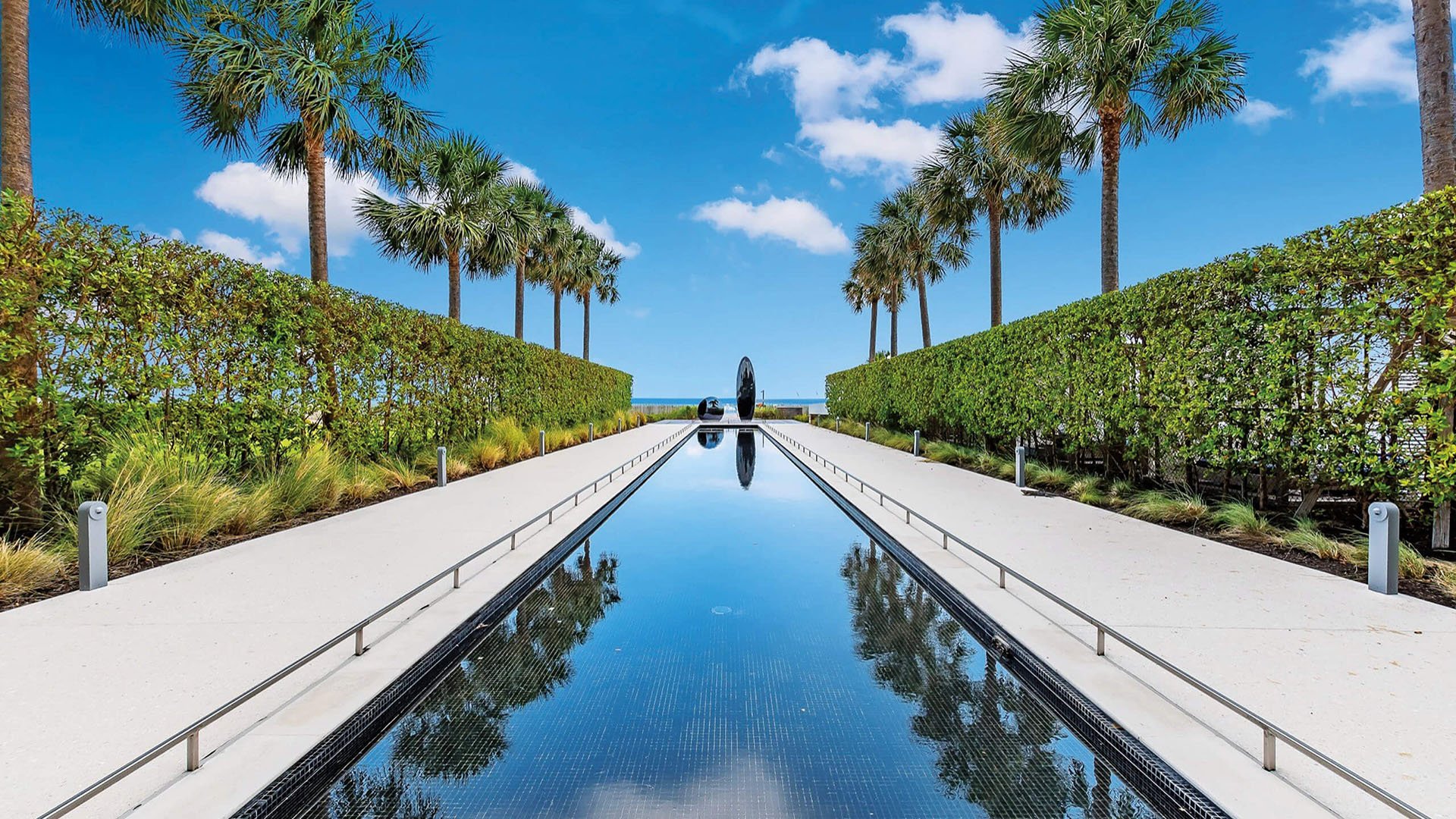 Palm-lined reflecting pool entry at Oceana Key Biscayne in Key Biscayne, showcasing luxury and ultra luxury condos with manicured hedges, sculpture, and a long water feature leading toward the ocean.