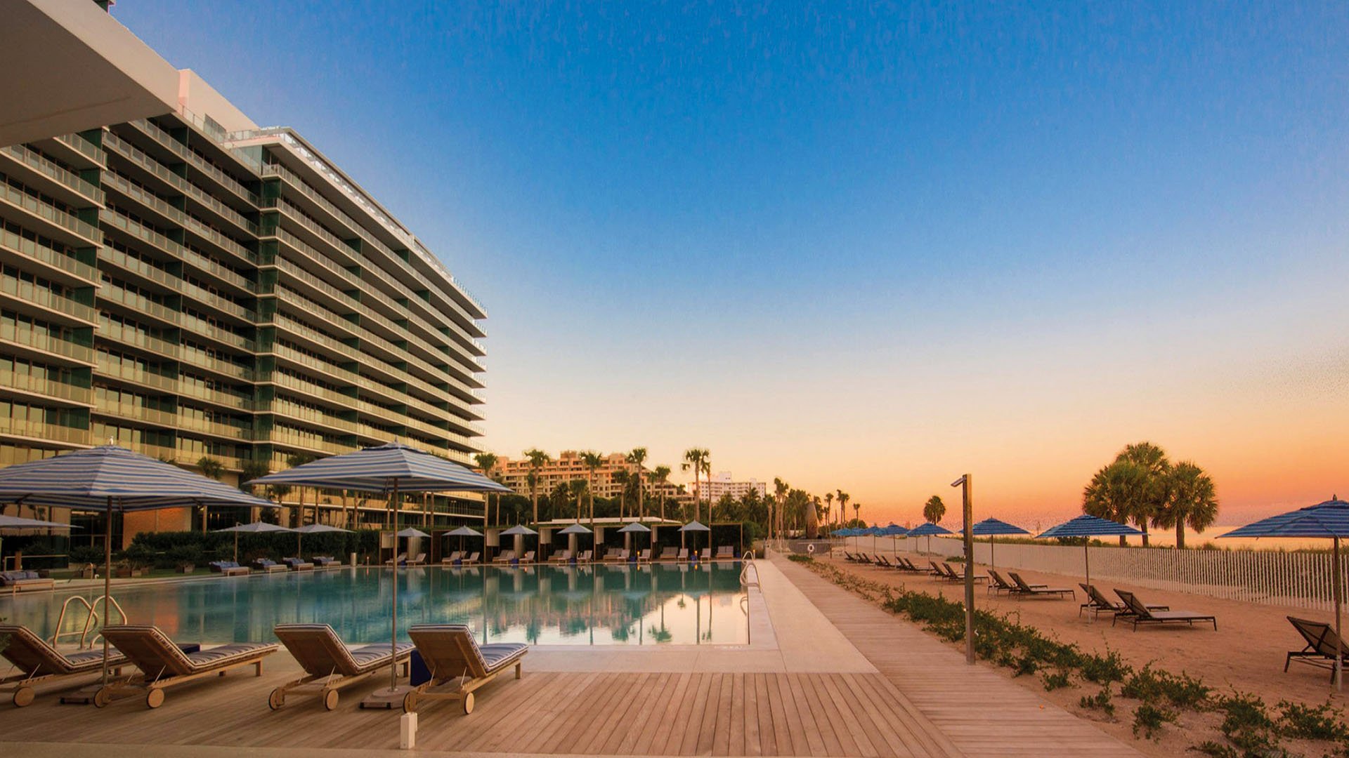 Sunset pool terrace at Oceana Key Biscayne in Key Biscayne, showing luxury and ultra luxury condos beside a beachfront pool deck with striped umbrellas, loungers, palms, and a glowing evening sky.
