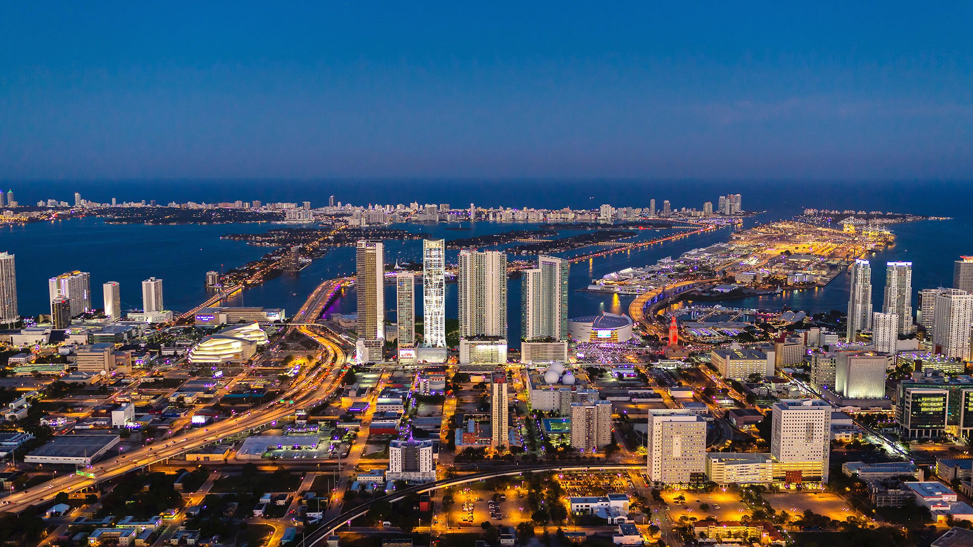 Twilight aerial from One Thousand Museum in Downtown Miami overlooking bridges, water, and the illuminated skyline, a view from luxury and ultra luxury condos.