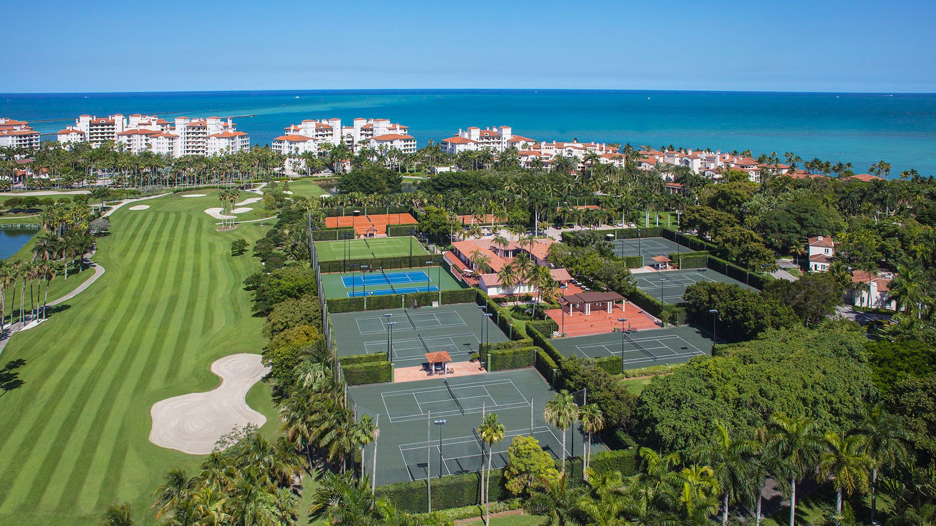 Aerial grounds view at Palazzo del Sol, Fisher Island, showing luxury and ultra luxury condos near tennis courts, manicured fairways, tropical landscaping, and the ocean horizon.