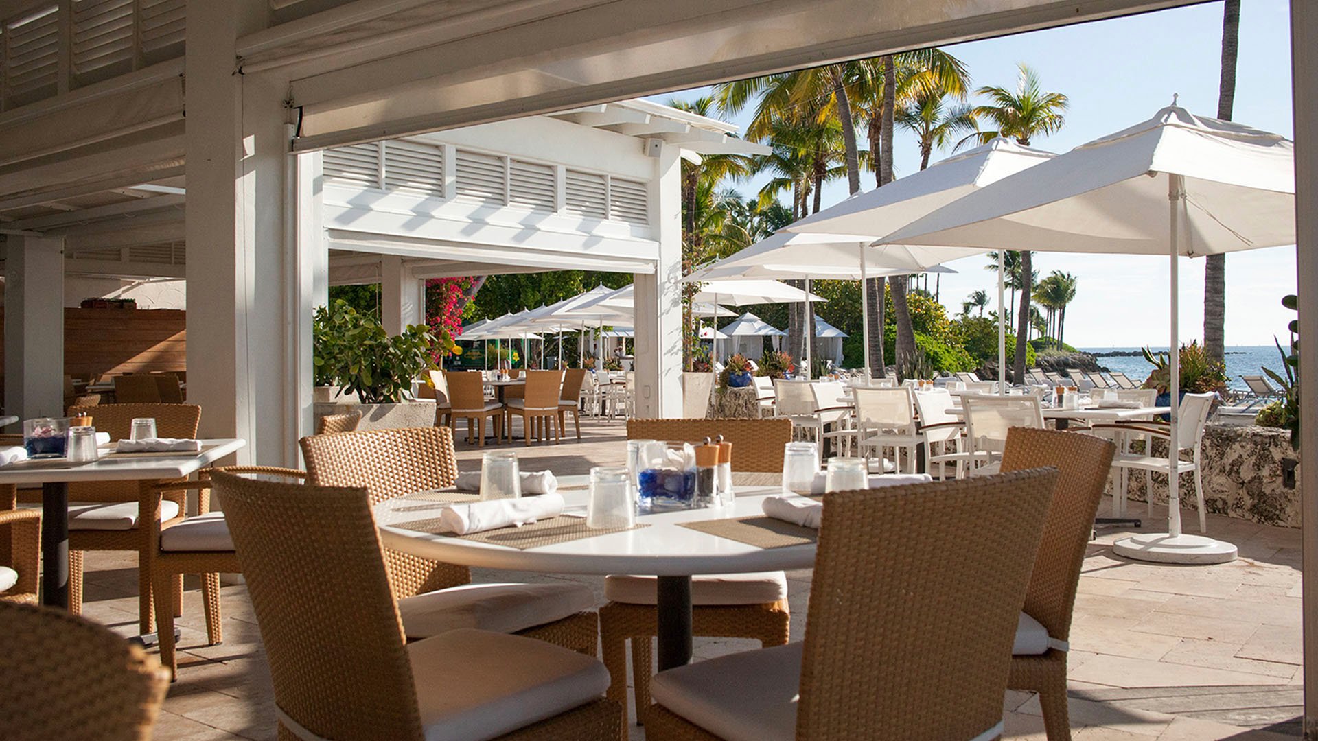 Beachfront dining terrace at Palazzo del Sol, Fisher Island, showing luxury and ultra luxury condos with cushioned seating, pergola shade, white umbrellas, and a relaxed resort atmosphere.