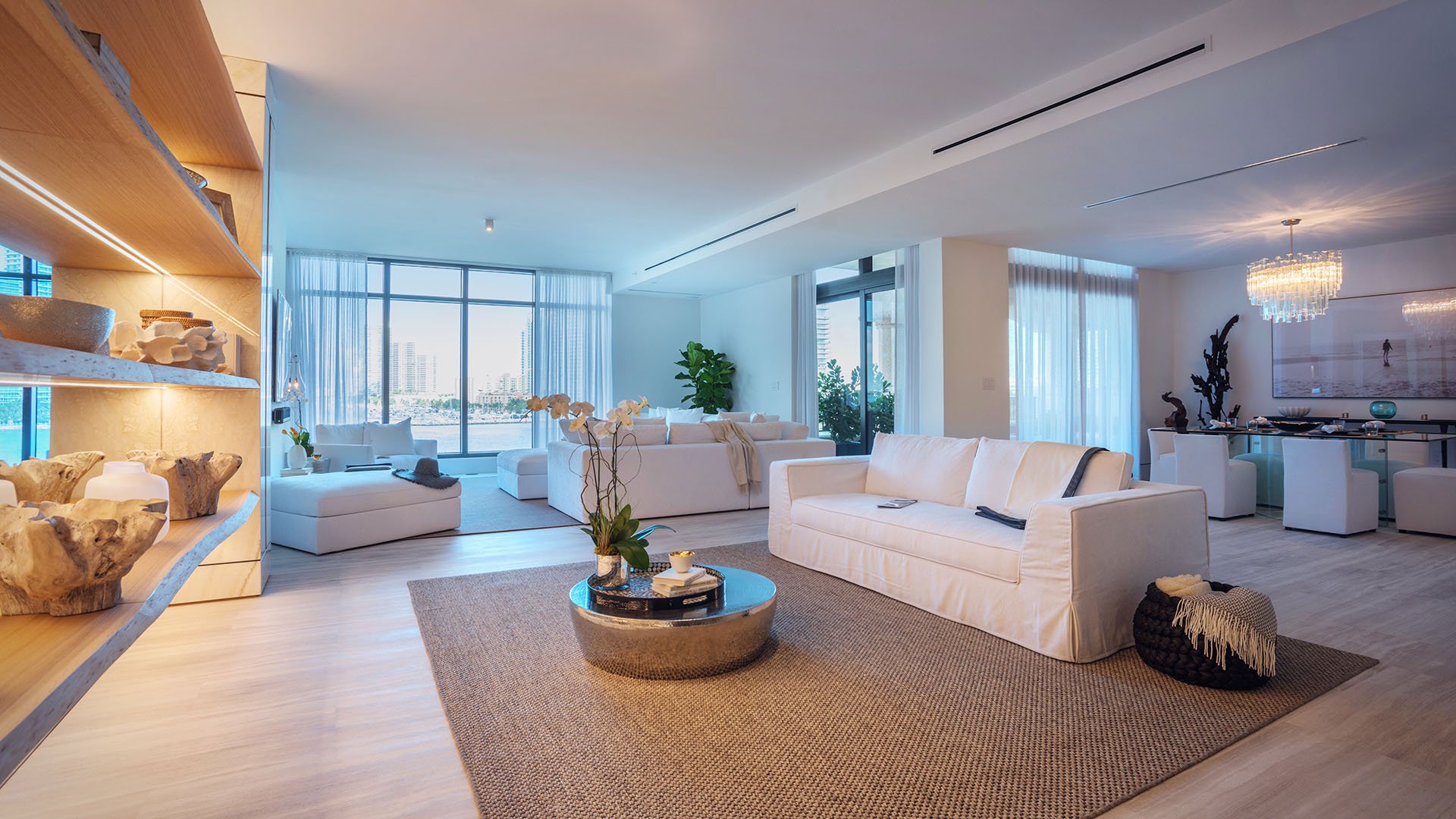 Bright waterfront living room at Palazzo del Sol, Fisher Island, showcasing luxury and ultra luxury condos with illuminated display shelving, pale wood floors, white seating, and an open kitchen.