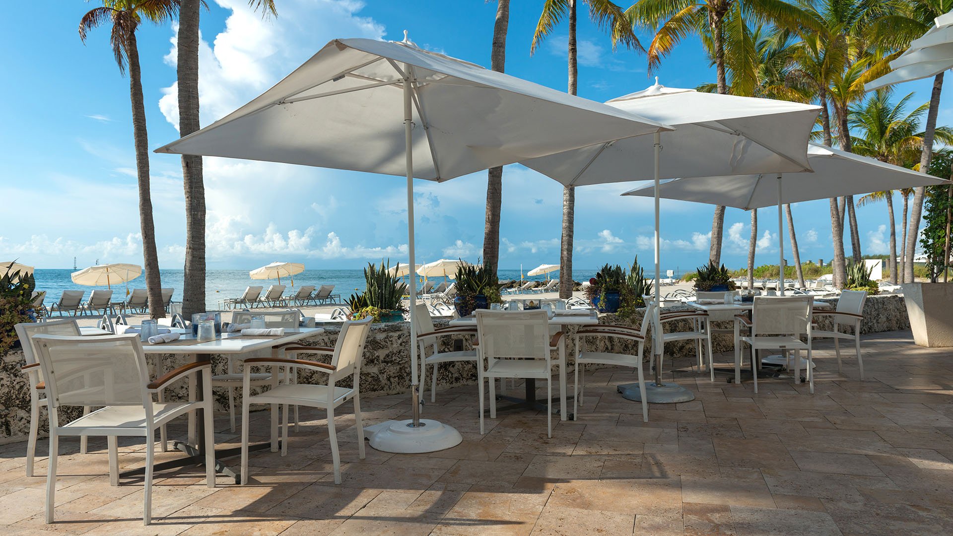 Oceanview dining terrace at Palazzo del Sol, Fisher Island, showing luxury and ultra luxury condos with white umbrellas, palm trees, and open-air tables set beside the beach.