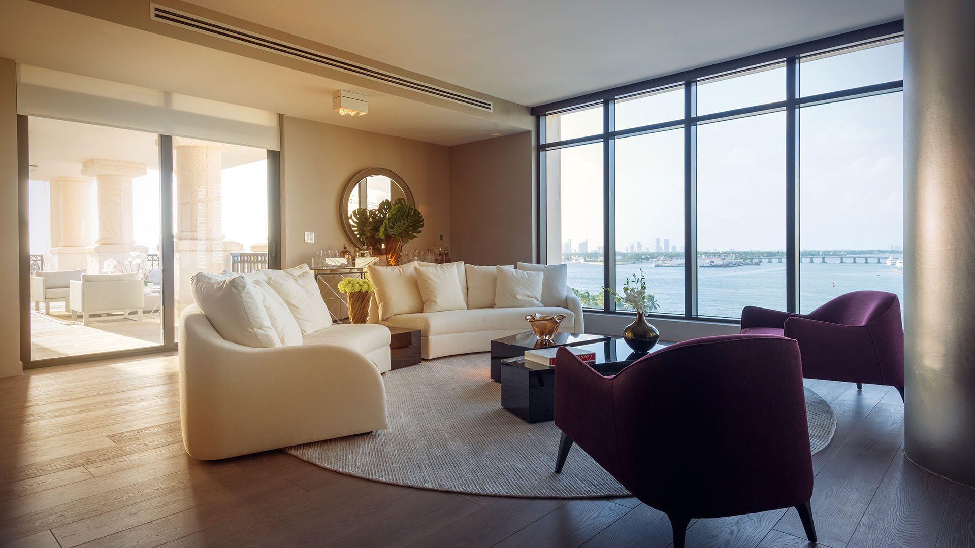 Sunlit corner living room at Palazzo del Sol, Fisher Island, highlighting luxury and ultra luxury condos with wraparound waterfront windows, cream seating, and warm late-day light.