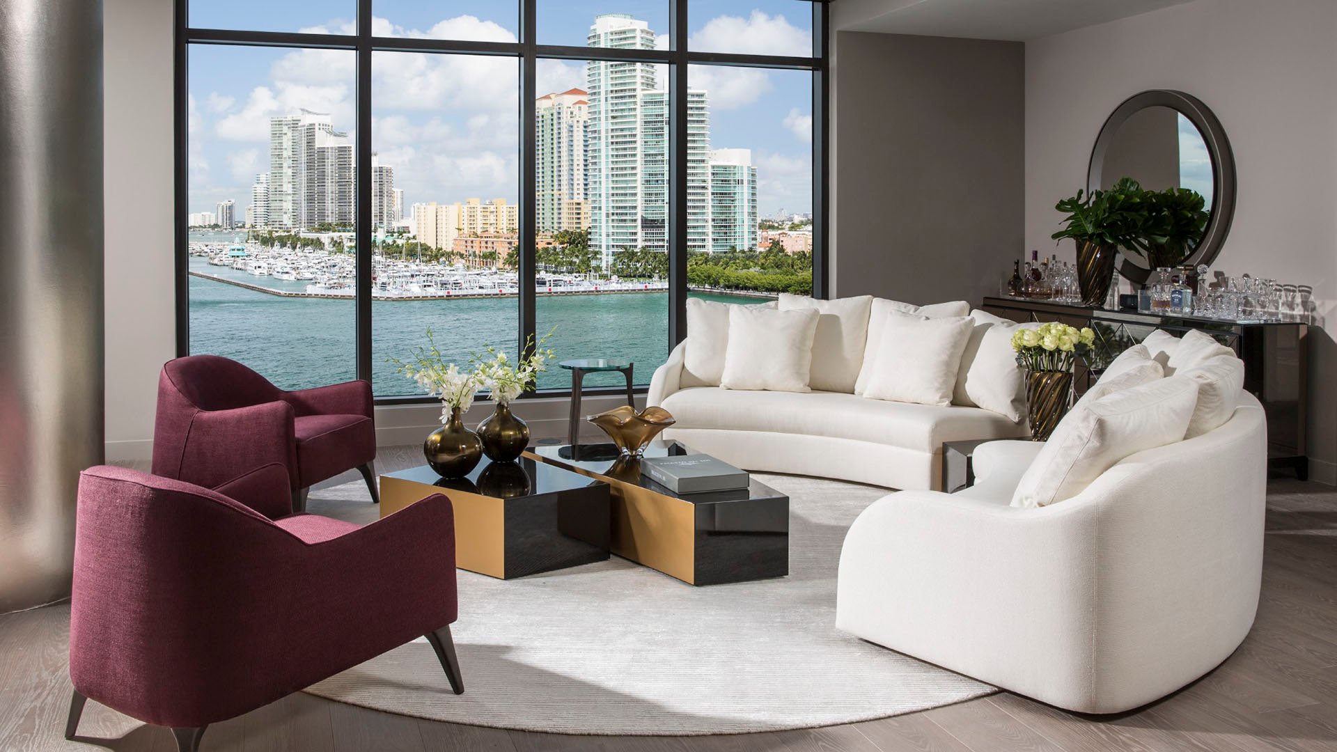 Waterfront sitting room at Palazzo del Sol, Fisher Island, showing luxury and ultra luxury condos with plum accent chairs, a creamy sofa, and close city and marina views through tall glass.