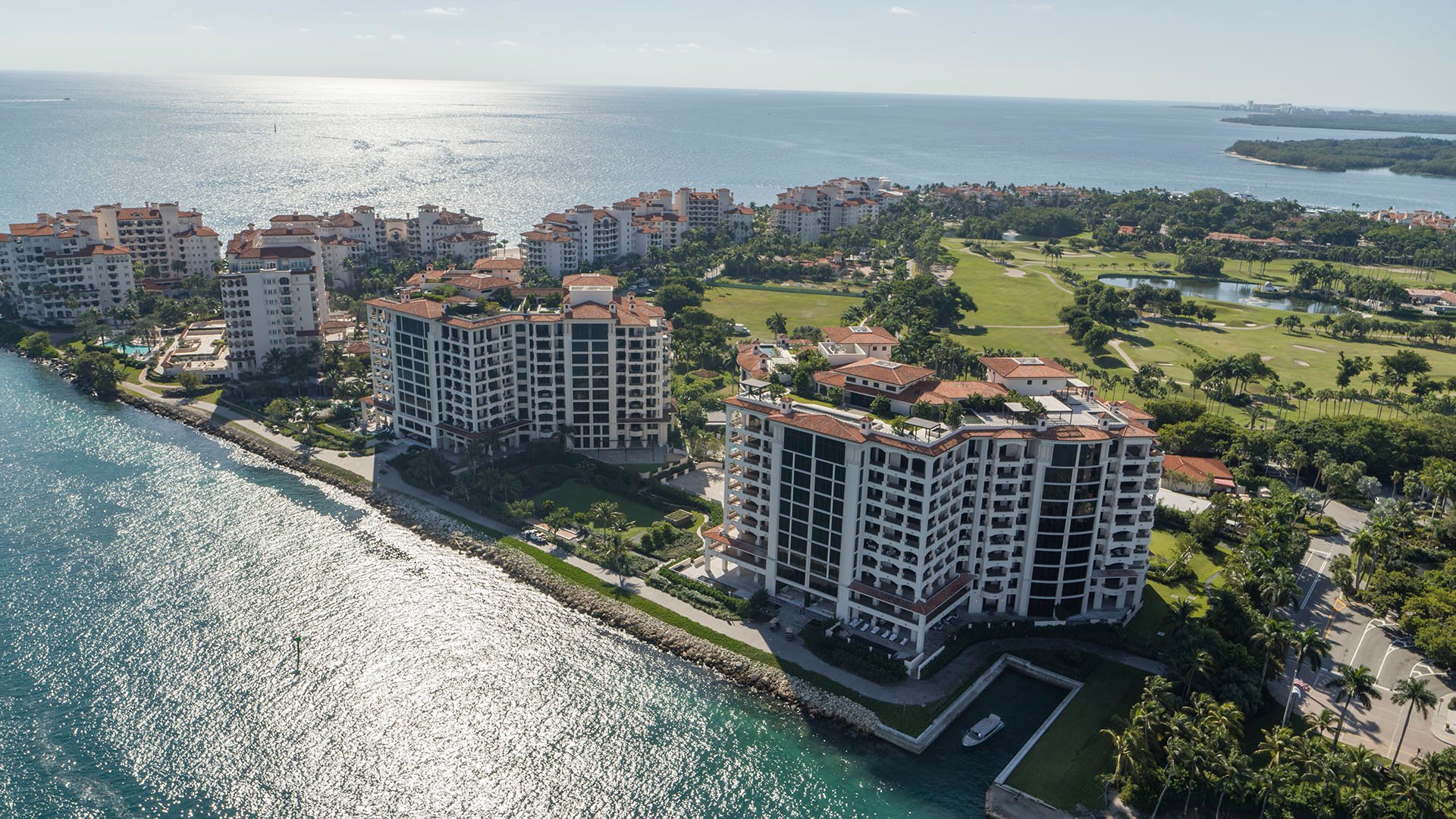 Palazzo della Luna in Fisher Island luxury and ultra luxury condos in an aerial waterfront view with sparkling shoreline water and a golf course beyond.