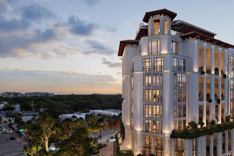 Ponce Park Residences Coral Gables, Miami twilight aerial of corner facade with lit windows and rooftop pergolas, highlighting luxury and ultra luxury preconstruction condos overlooking tree-lined city views.