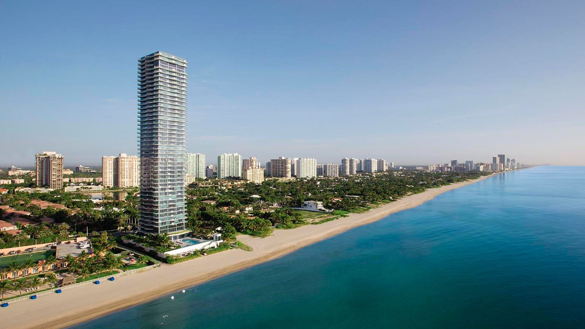 Aerial beachfront panorama of Regalia in Sunny Isles Beach showing luxury and ultra luxury condos along a quiet stretch of sand and blue ocean.