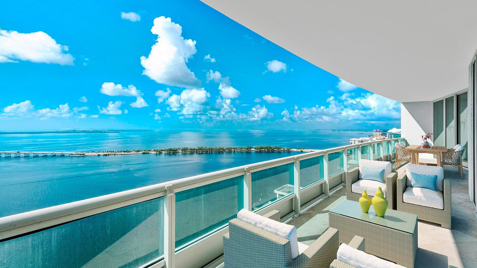 Panoramic balcony terrace at Santa Maria in Brickell, featuring luxury and ultra luxury condos with outdoor lounge seating, glass railings, and wide waterfront views beneath a curved overhang.
