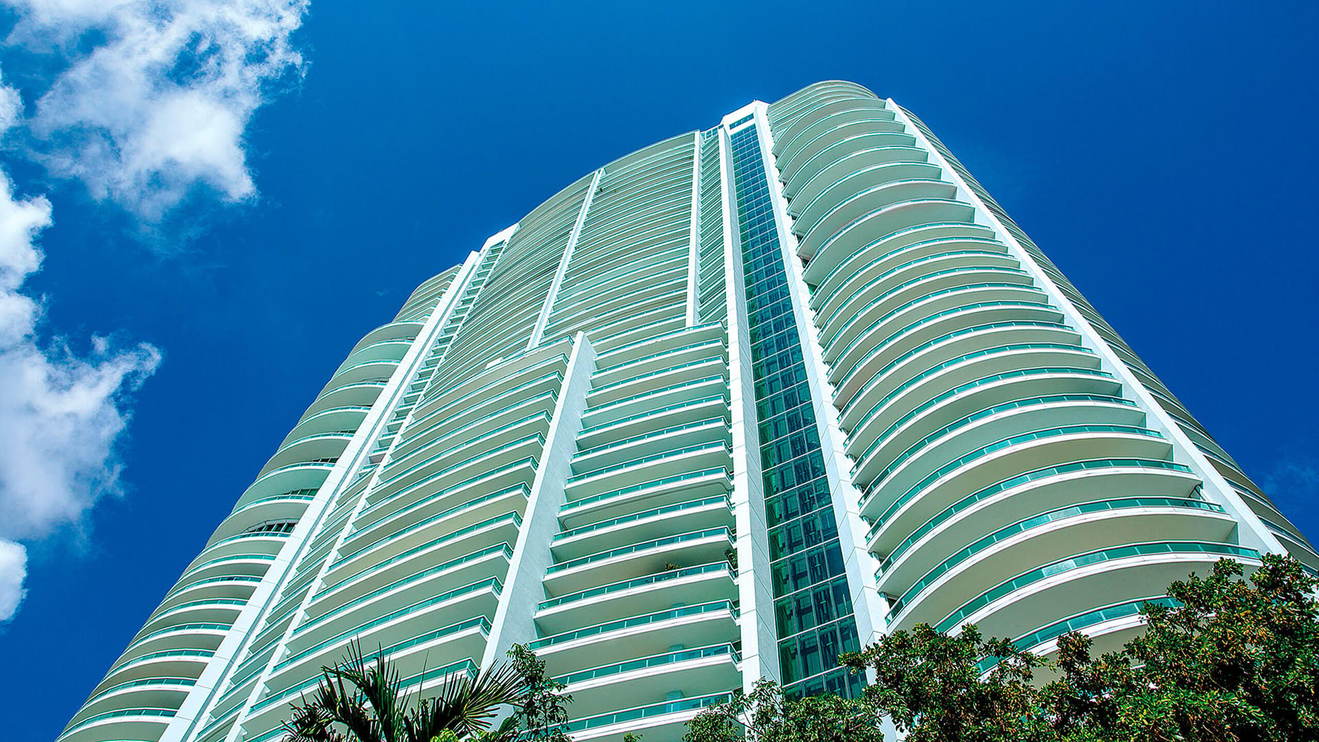 Skyward tower view of Santa Maria in Brickell, capturing luxury and ultra luxury condos with expansive curved balconies, a striking vertical glass core, and vivid blue sky above.