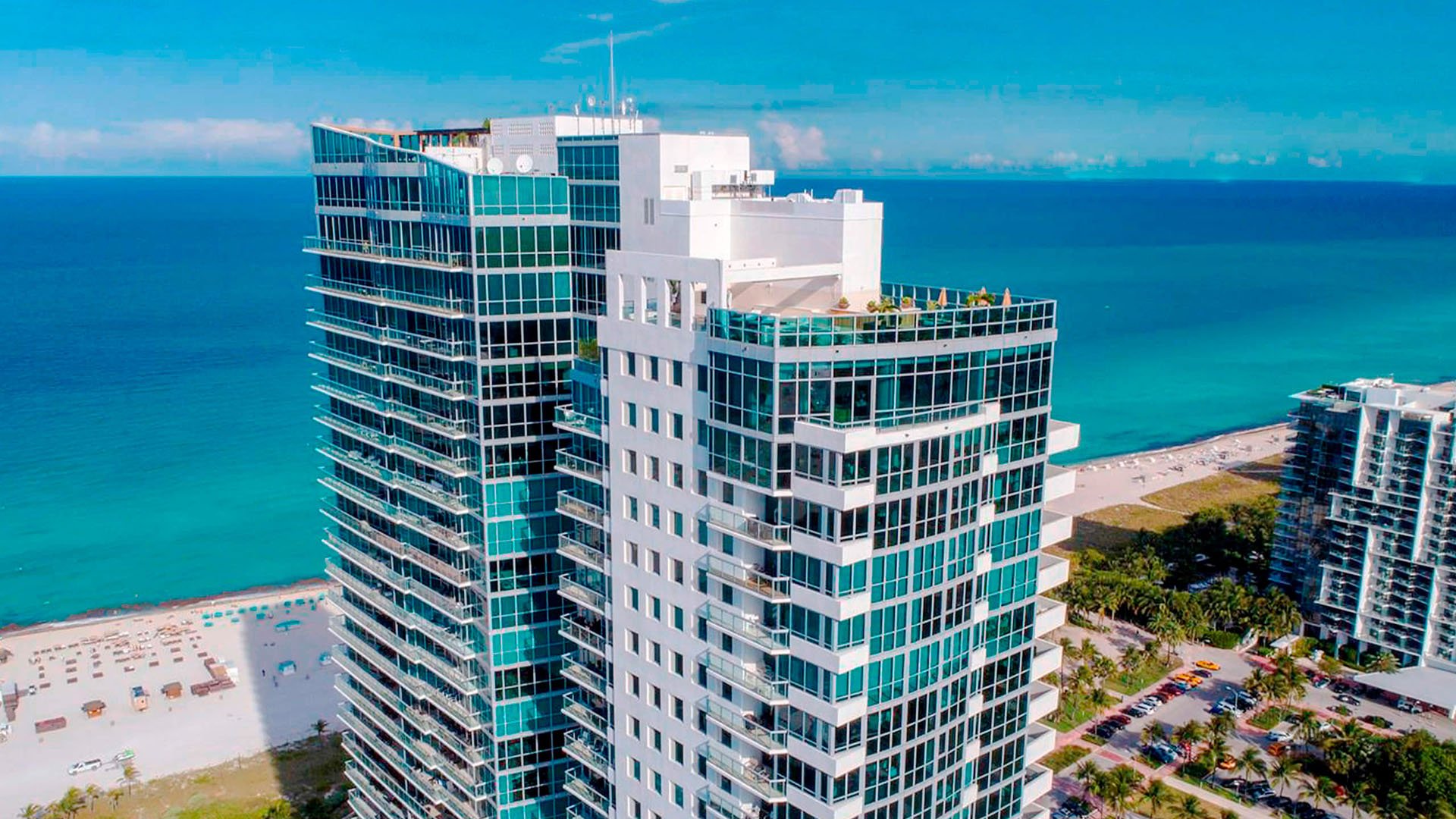 Aerial waterfront view of Setai Miami Beach in Miami Beach featuring luxury and ultra luxury condos, a beachfront glass tower, turquoise Atlantic Ocean, and sandy shoreline.