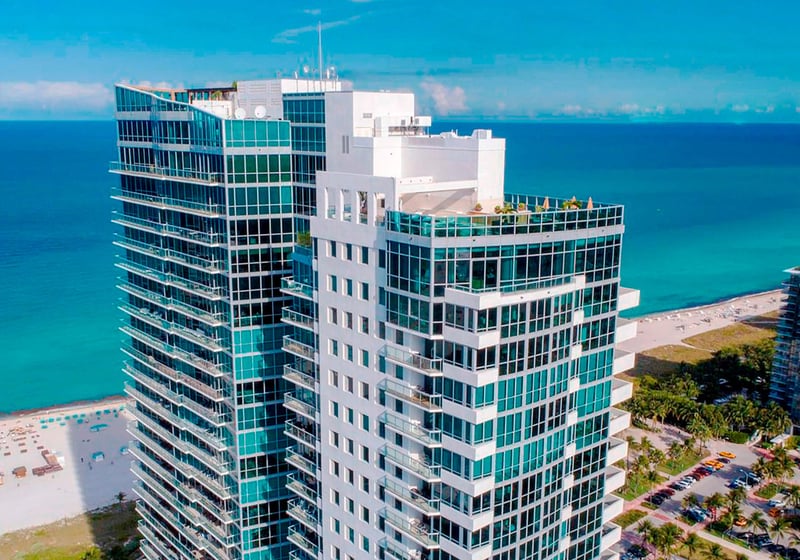 Aerial waterfront view of Setai Miami Beach in Miami Beach featuring luxury and ultra luxury condos, a beachfront glass tower, turquoise Atlantic Ocean, and sandy shoreline.