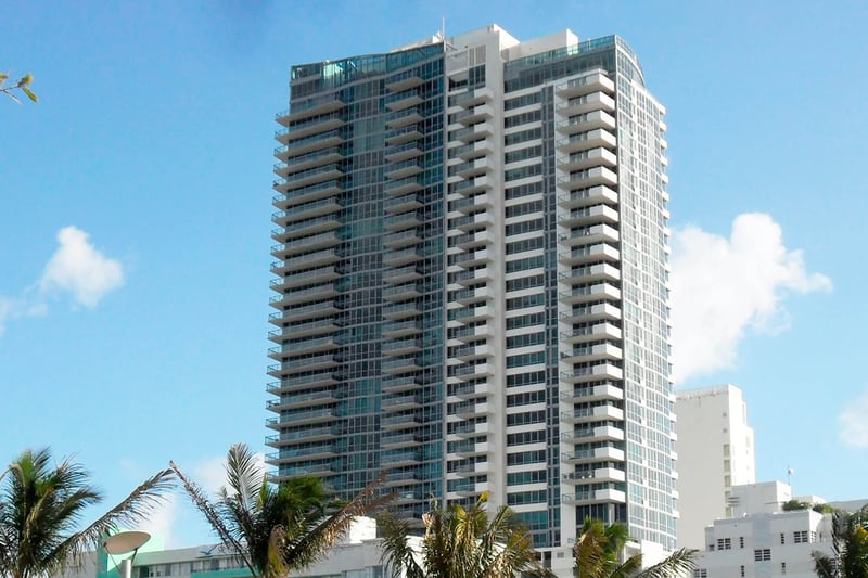 Exterior view of Setai Miami Beach in Miami Beach showing luxury and ultra luxury condos in a tall glass tower with wraparound balconies, palm trees, and a bright blue sky.