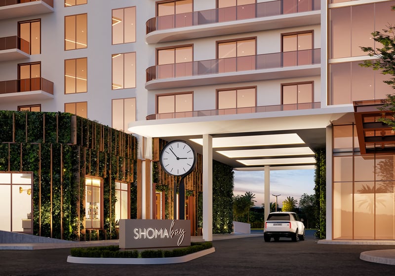 Shoma Bay North Bay Village, Miami, Florida porte-cochere entrance at dusk with illuminated driveway canopy, clock and branded signage, arriving to luxury and ultra luxury preconstruction condos in a mixed-use setting.