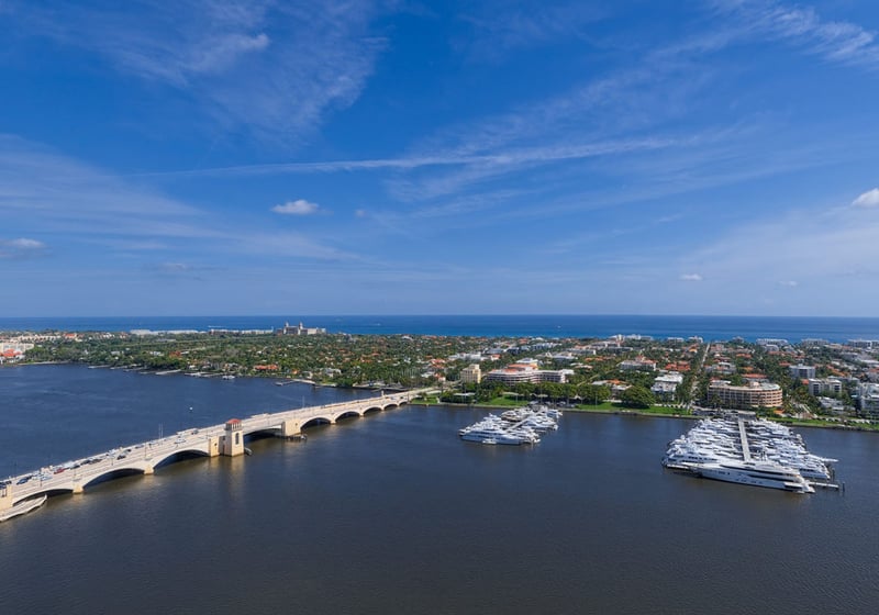 Aerial view of a bridge, yacht marina, and waterfront neighborhood near The Bristol Palm Beach in Palm Beach, showcasing luxury and ultra luxury condos with expansive water and skyline vistas.