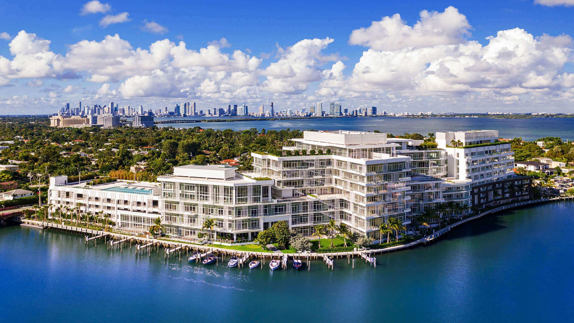 Aerial waterfront overview with marina slips and a distant skyline at The Ritz-Carlton Residences Miami Beach in Miami Beach, presenting luxury and ultra luxury condos on a broad bayfront site.