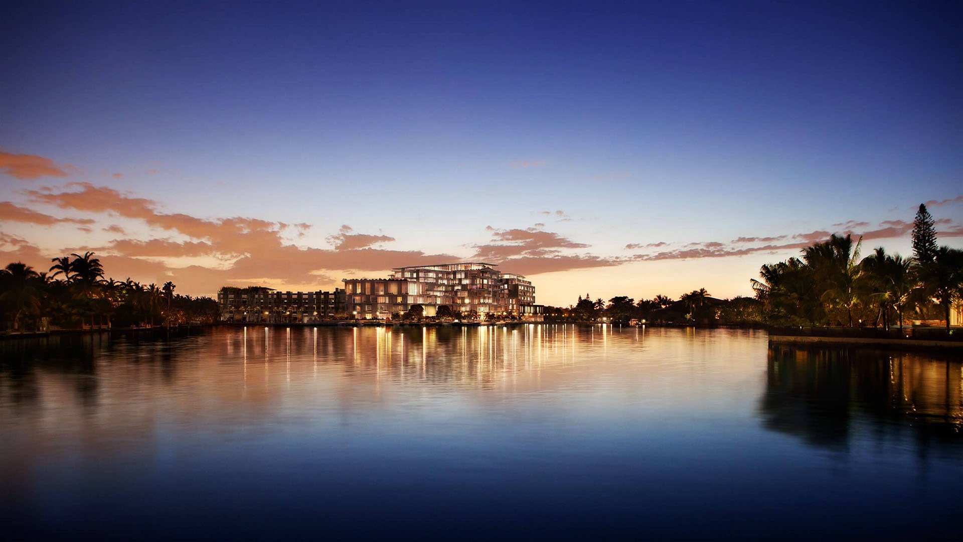 Twilight waterfront exterior with glowing reflections at The Ritz-Carlton Residences Miami Beach in Miami Beach, showing luxury and ultra luxury condos across calm water framed by palms.