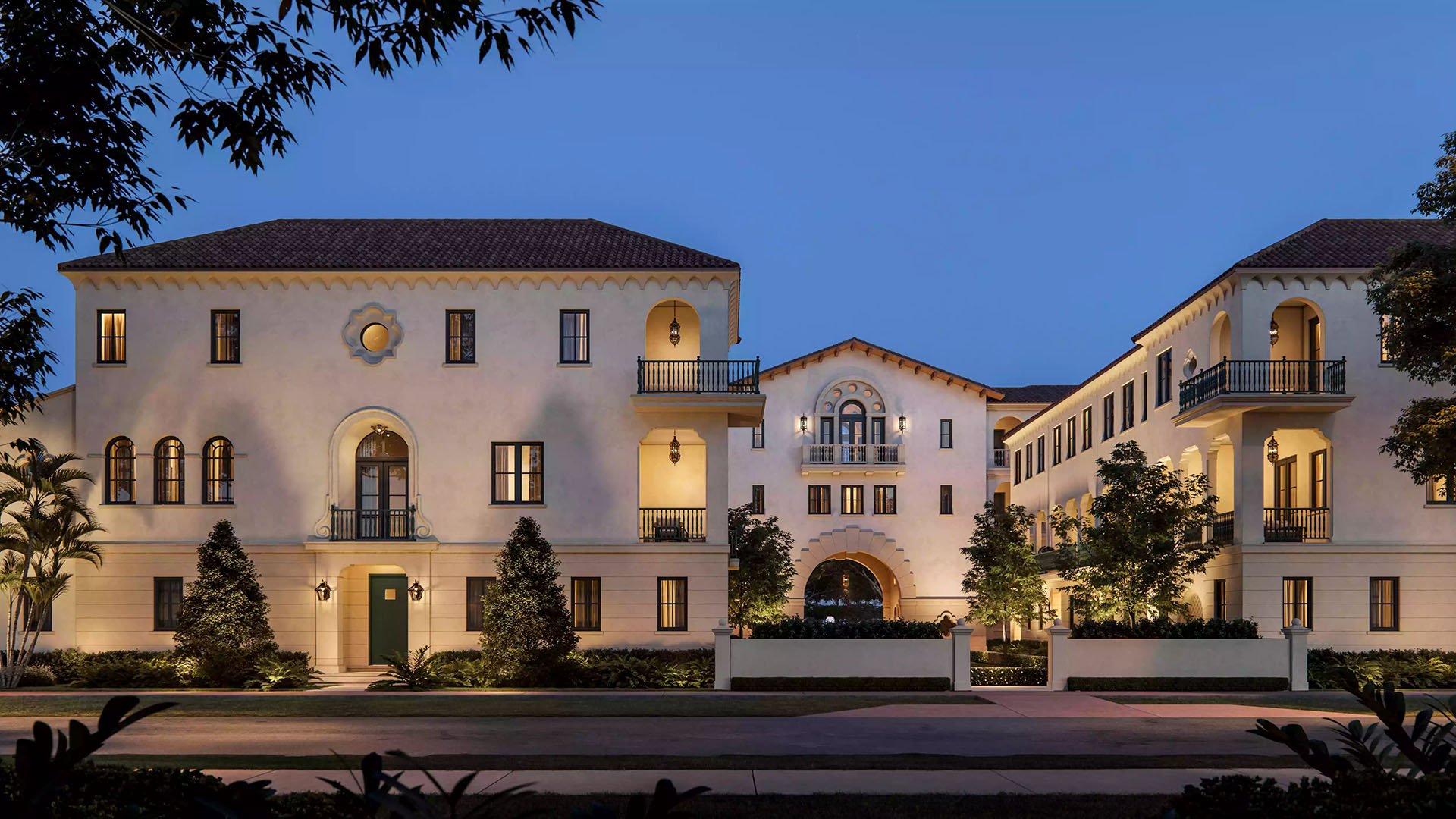 The Village at Coral Gables in Coral Gables, Miami dusk street view with illuminated archway, lanterns and Spanish Mediterranean balconies; luxury and ultra luxury preconstruction condos at twilight.
