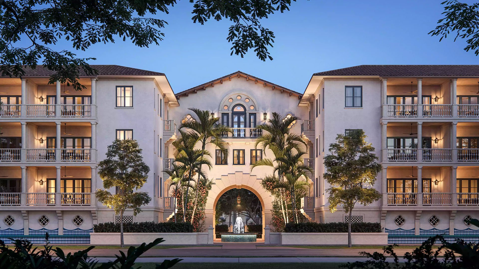 The Village at Coral Gables in Coral Gables, Miami twilight view of central archway, palm trees and courtyard fountain lit for evening; luxury and ultra luxury preconstruction condos residences.