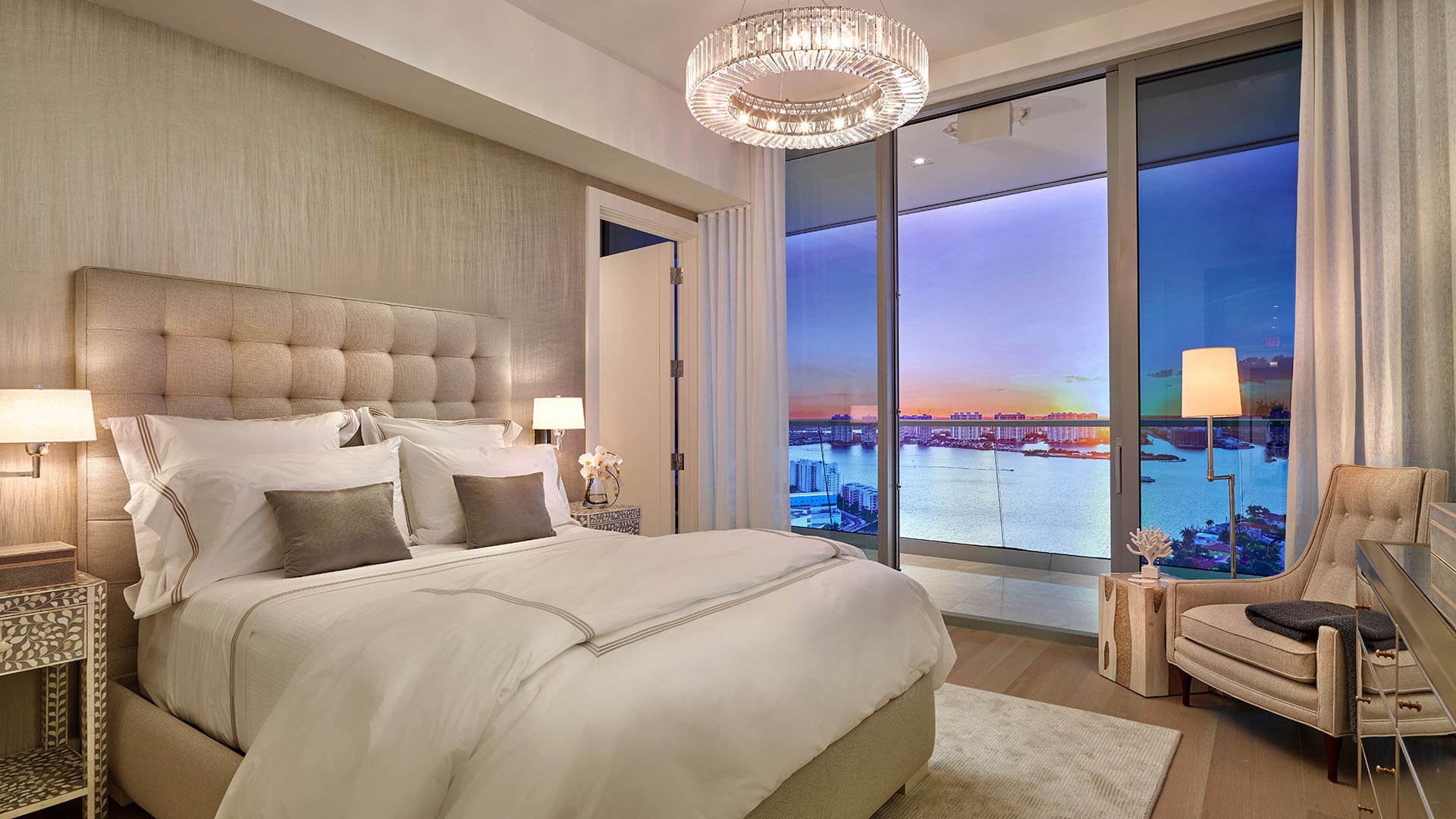 Turnberry Ocean Club in Sunny Isles Beach luxury and ultra luxury condos showcase a bedroom with a tufted headboard, chandelier, balcony doors, and sunset water views.