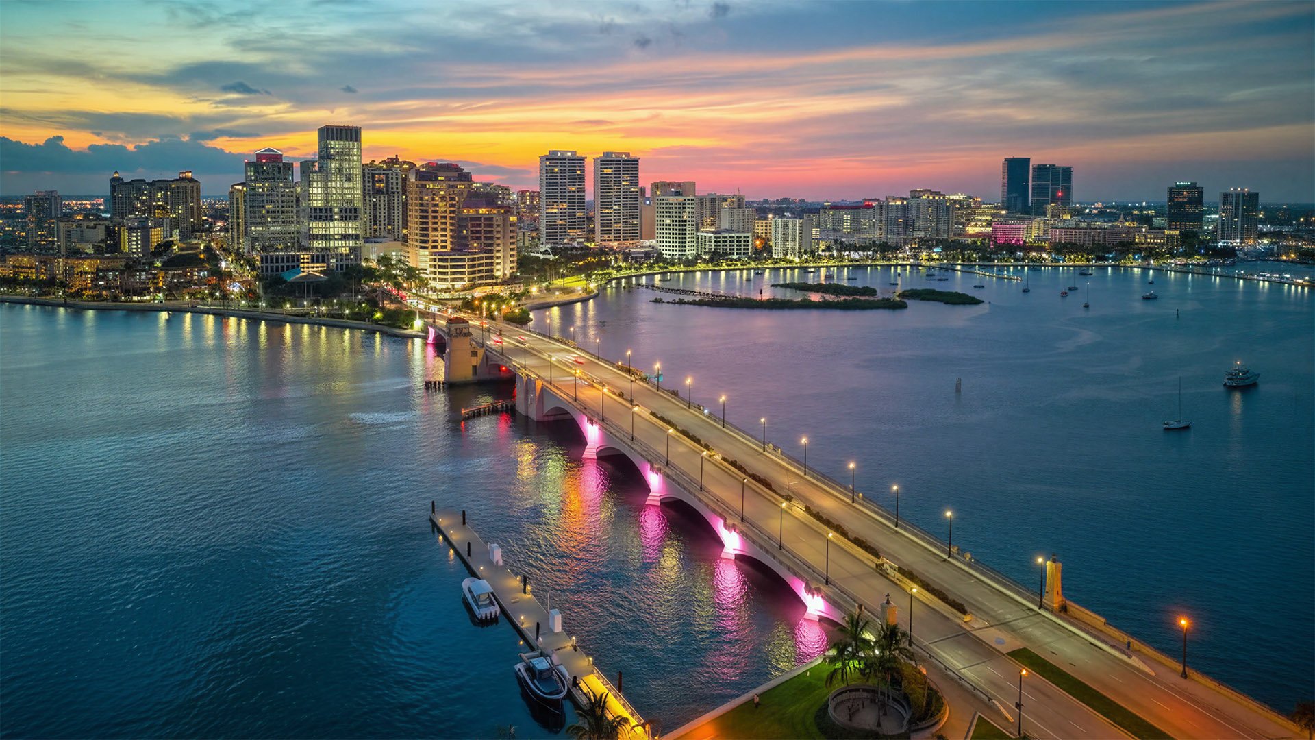 West Palm Beach luxury and ultra luxury condos in an aerial waterfront skyline view at sunset with an illuminated bridge over the Intracoastal, downtown high-rise residences, city lights, small islands, and yachts on calm water.