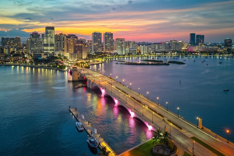 West Palm Beach luxury and ultra luxury condos in an aerial waterfront skyline view at sunset with an illuminated bridge over the Intracoastal, downtown high-rise residences, city lights, small islands, and yachts on calm water.