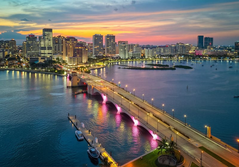 West Palm Beach luxury and ultra luxury condos in an aerial waterfront skyline view at sunset with an illuminated bridge over the Intracoastal, downtown high-rise residences, city lights, small islands, and yachts on calm water.