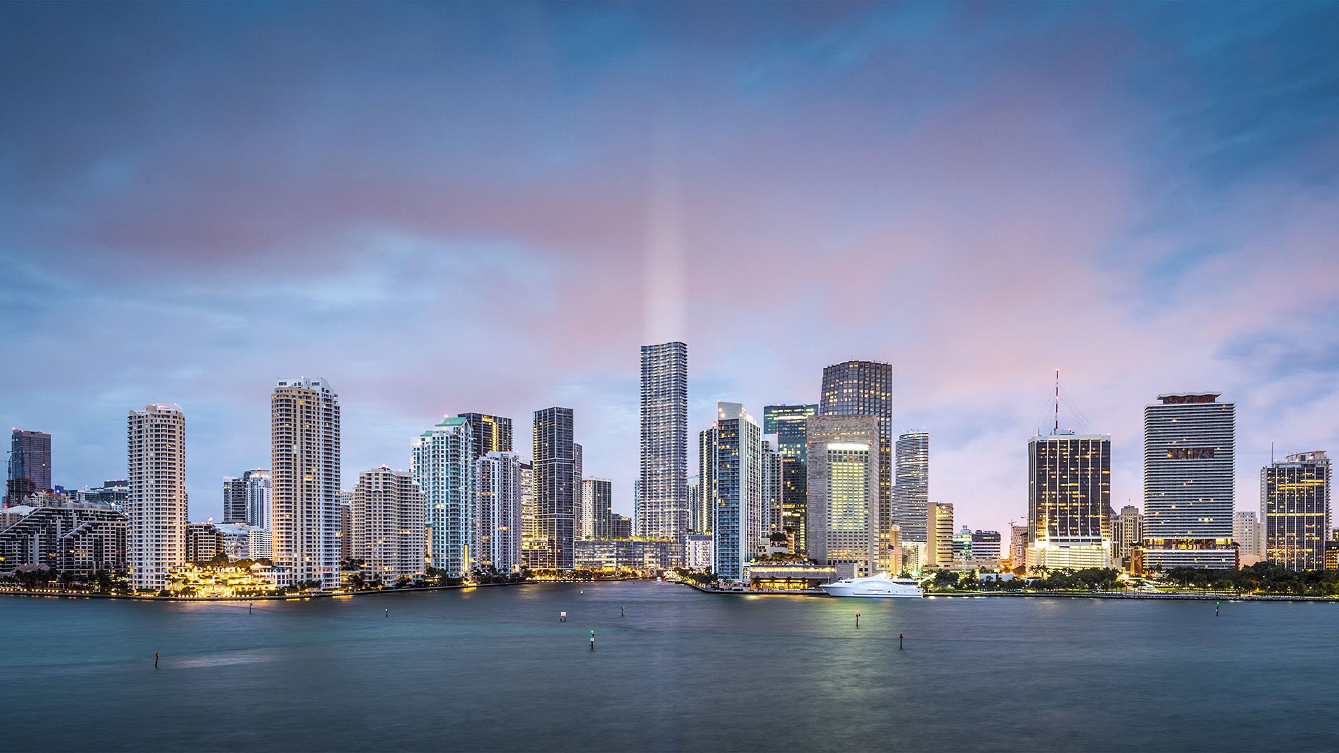 Baccarat Residences in Brickell, Miami, luxury and ultra luxury condos featuring an evening panorama of the waterfront skyline, illuminated towers, soft clouds, and calm bay water in the foreground.
