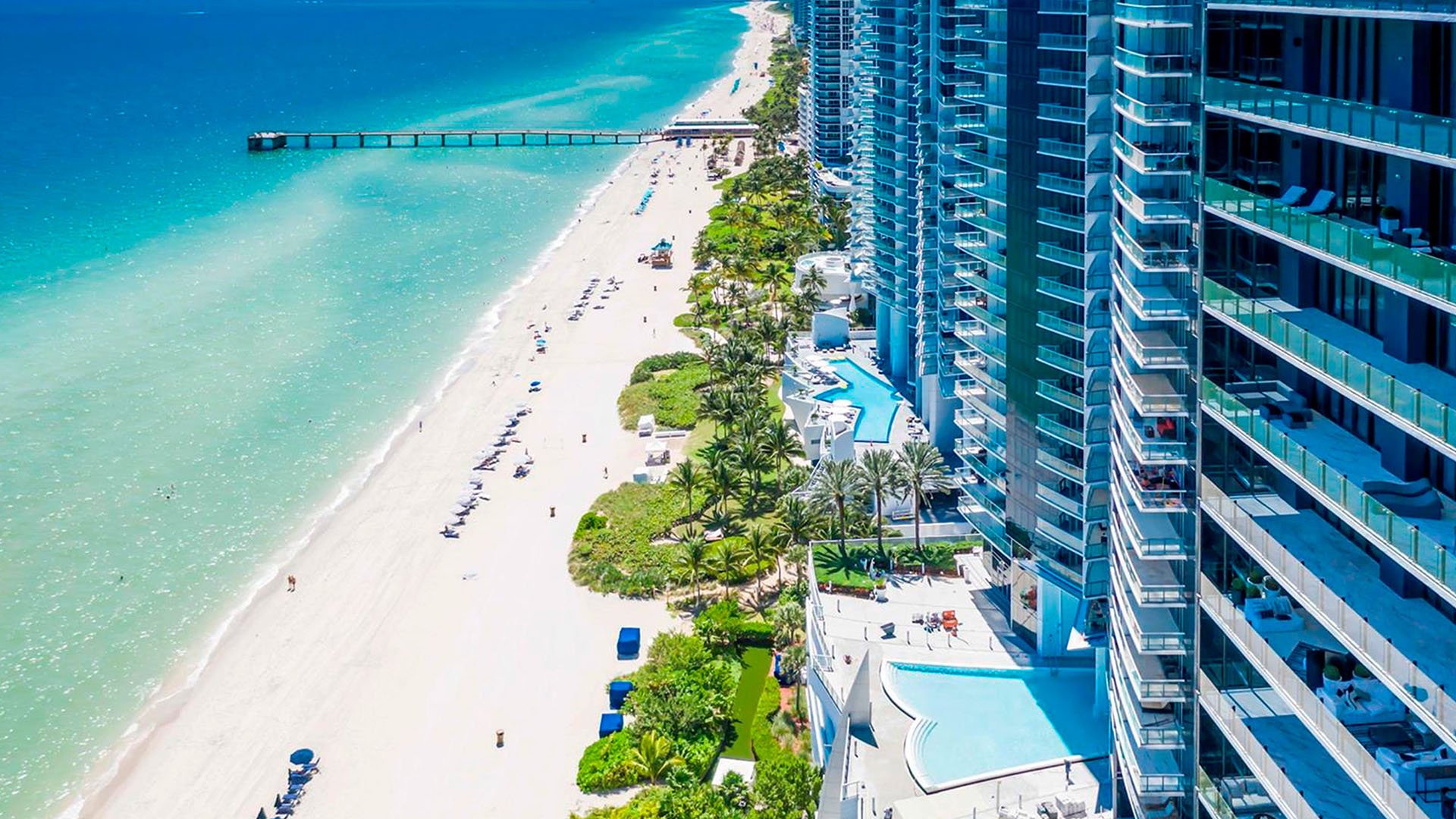 Aerial beachfront skyline view of Jade Ocean in Sunny Isles Beach, showing luxury and ultra luxury condos along turquoise water with a long pier, sandy shoreline, and neighboring oceanfront towers.